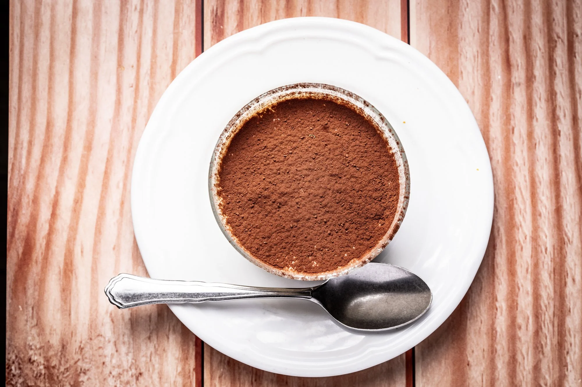 Un tiramisù ricoperto di cacao in polvere sopra un piatto bianco, con un cucchiaio accanto.