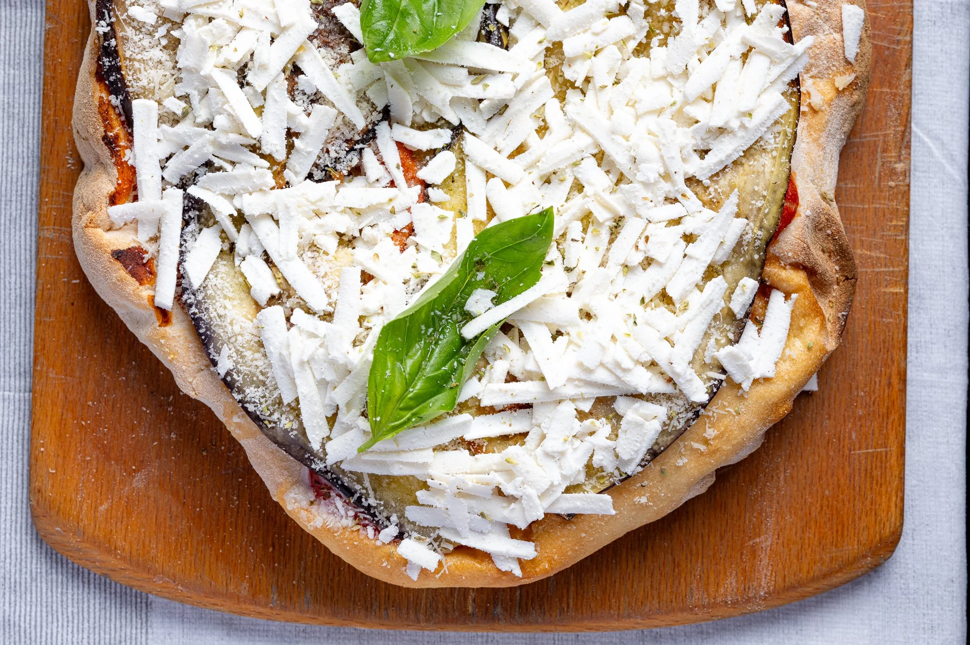 Pizza con ricotta salata, basilico e melanzane, su tagliere di legno.