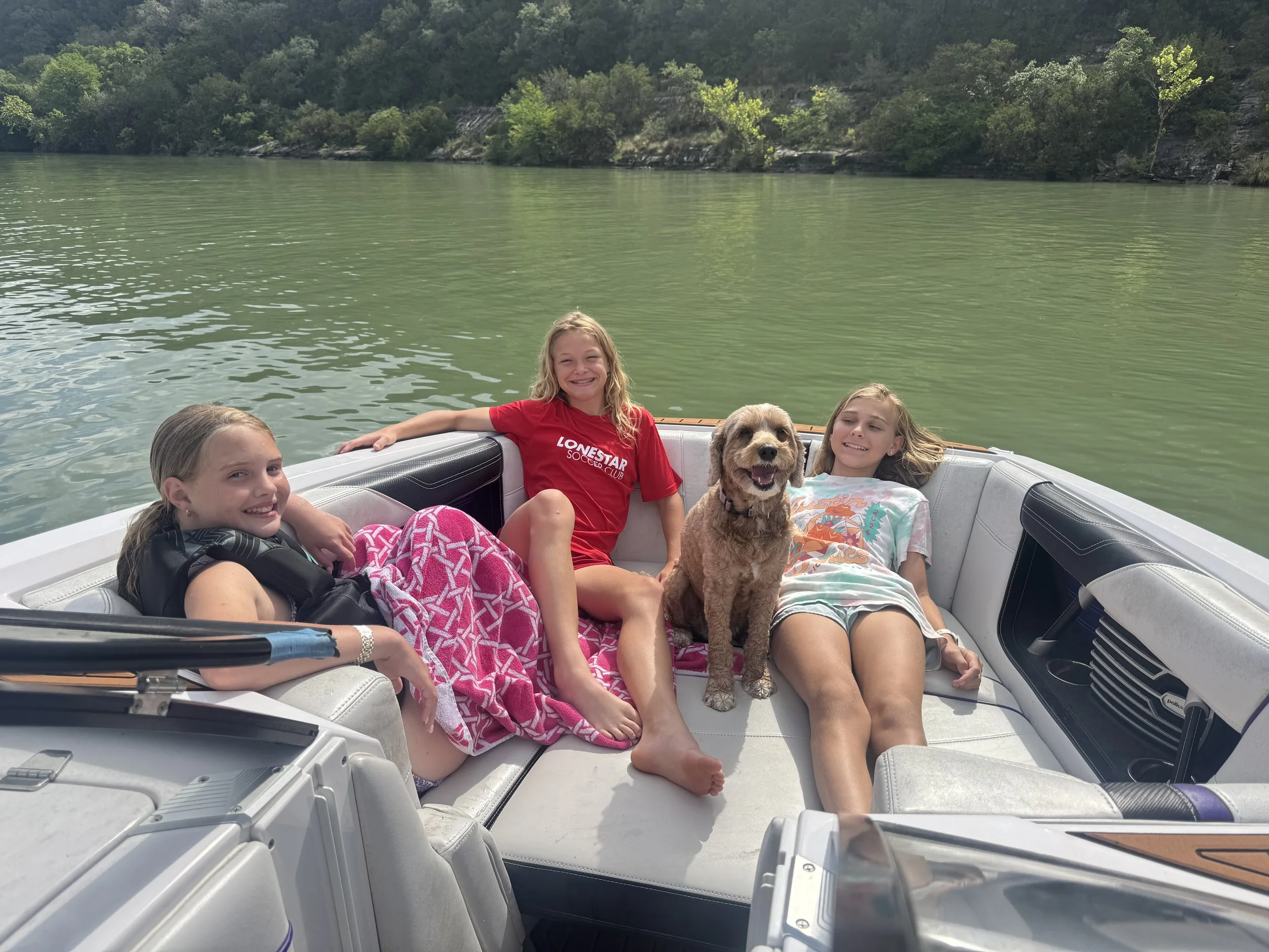 Three girls and a dog relaxing on a boat on a lake, with green water and trees on the shoreline in the background.