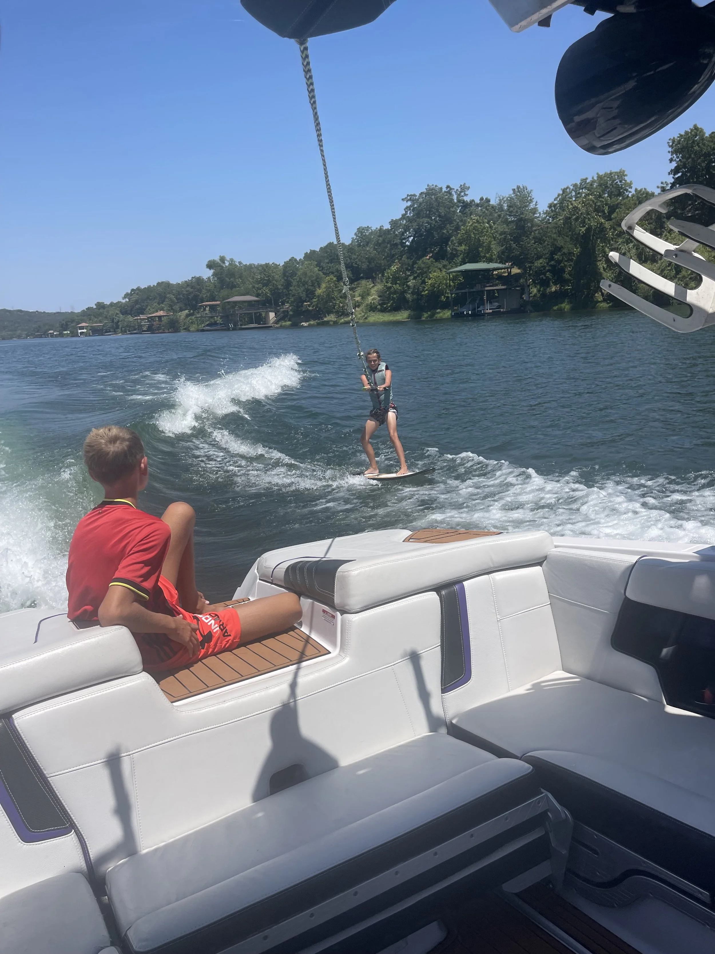 A person wakeboarding on a lake, being pulled by a boat, with another person relaxing on the boat's cushioned seats, and lush green trees in the background.