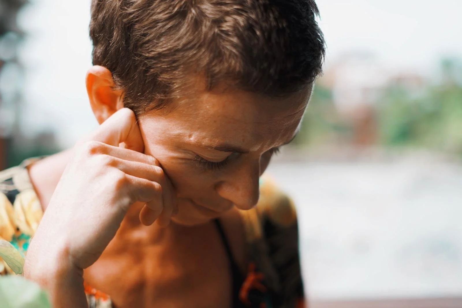 Persona joven con cabello corto, tocándose la oreja y con expresión pensativa, en un entorno exterior.