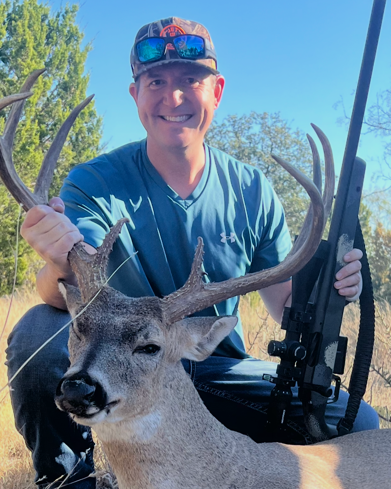 A man smiling outdoors in a blue shirt and camouflage cap, holding a rifle, poses with a large buck deer with antlers.