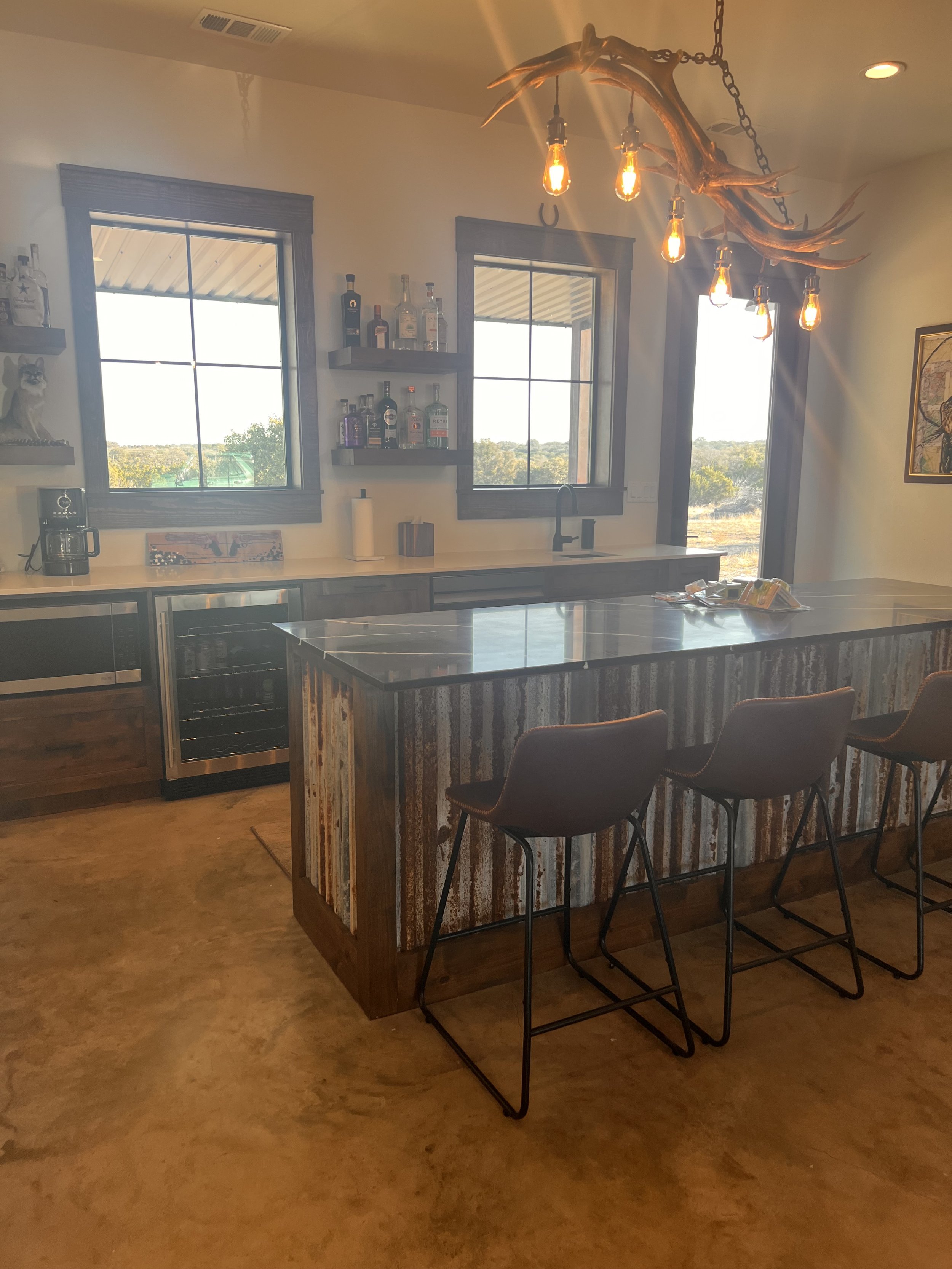 Rustic kitchen with two windows, a bar island with three bar stools, a countertop with bottles and a coffee machine, and a unique chandelier made of antlers with hanging Edison bulbs.