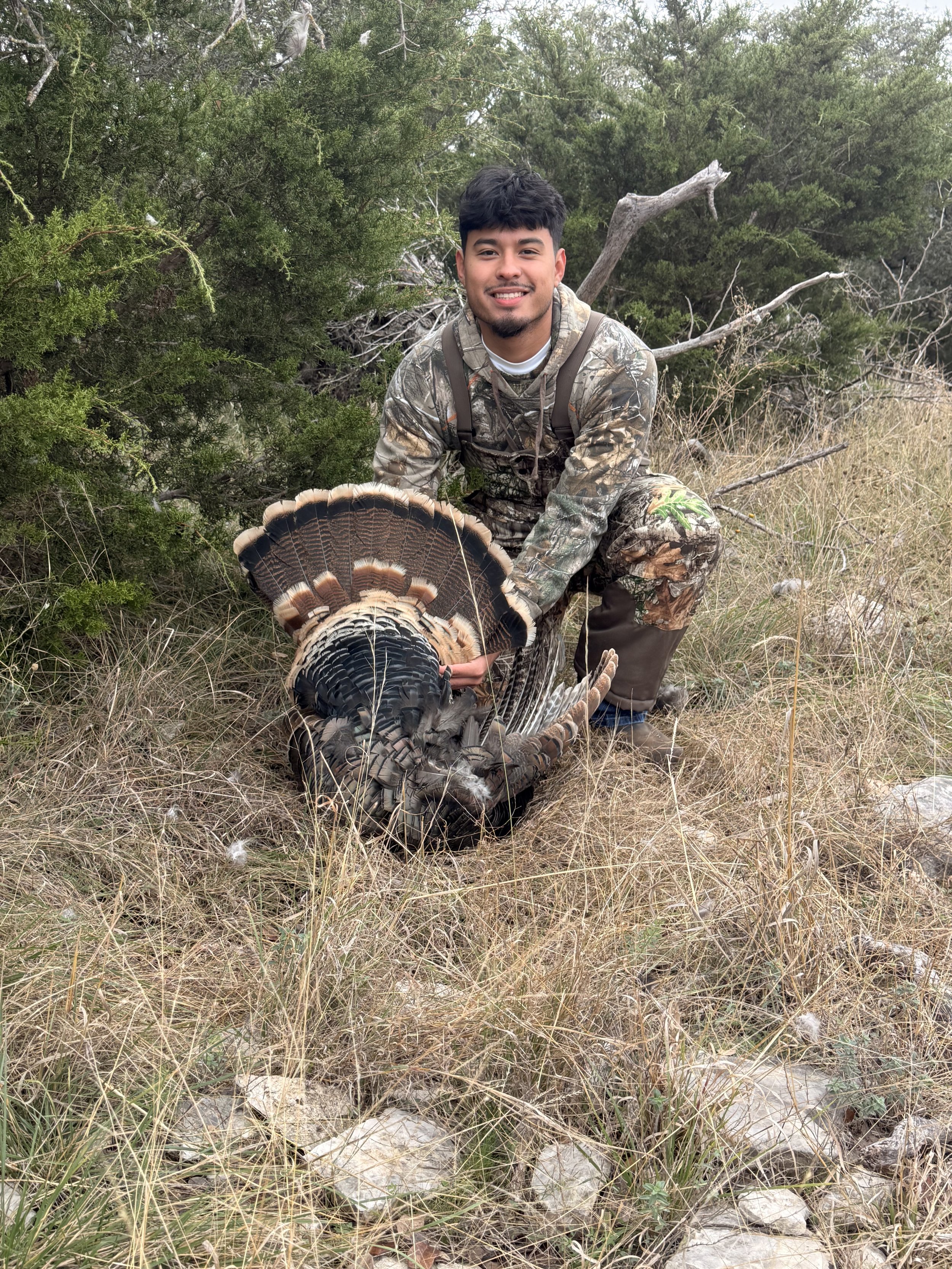 A young man in camouflage clothing kneels on dry grass in a wooded area, holding a large wild turkey with its feathers fanned out.