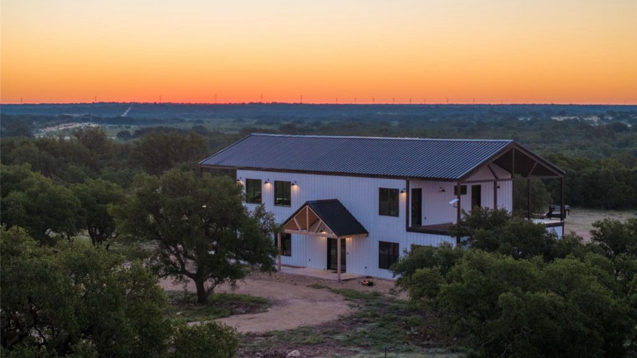 A two-story modern house with metal roof and white exterior, surrounded by trees, at sunset in a rural area.