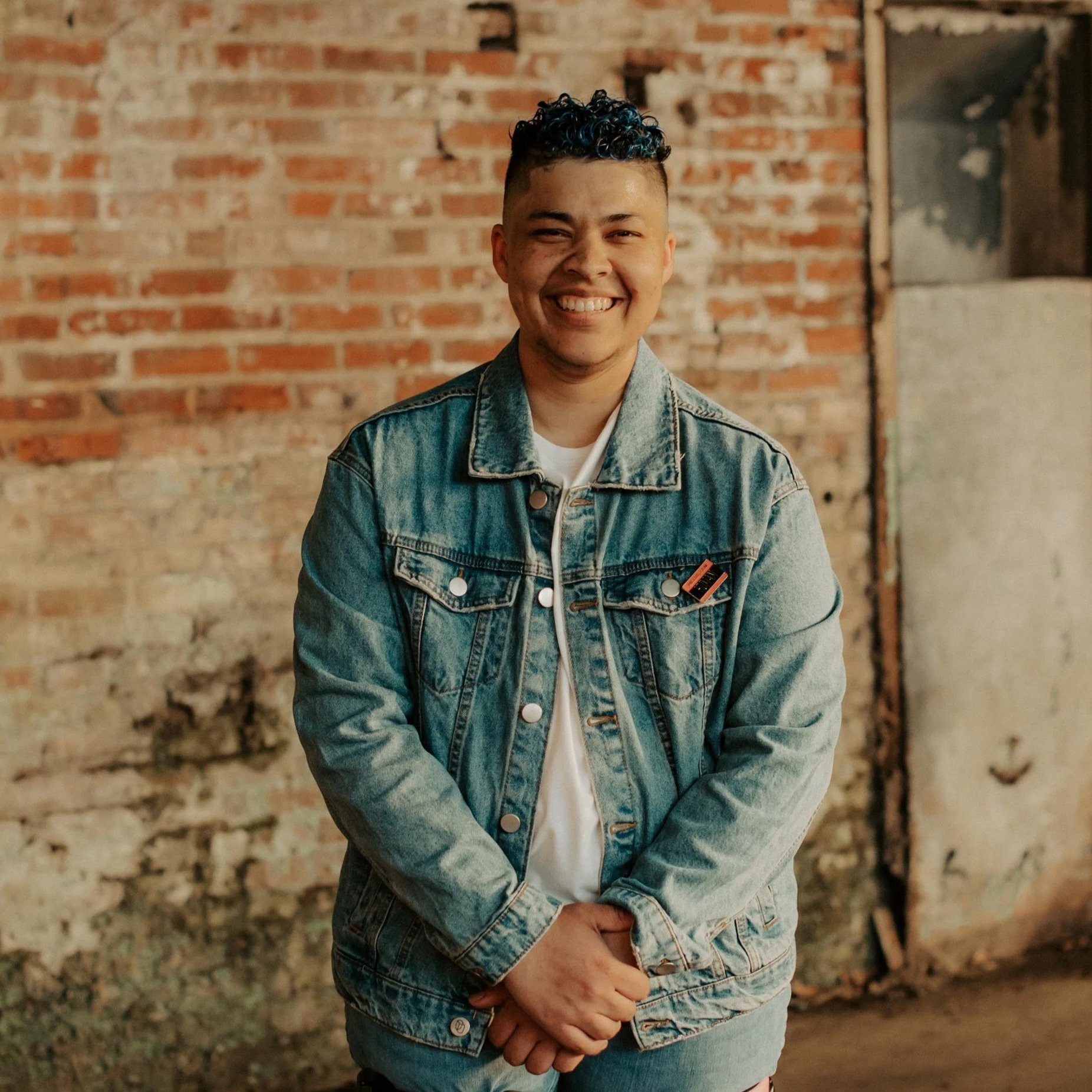 A smiling young man with curly black hair stands in front of a brick wall, wearing a denim jacket and a white t-shirt.