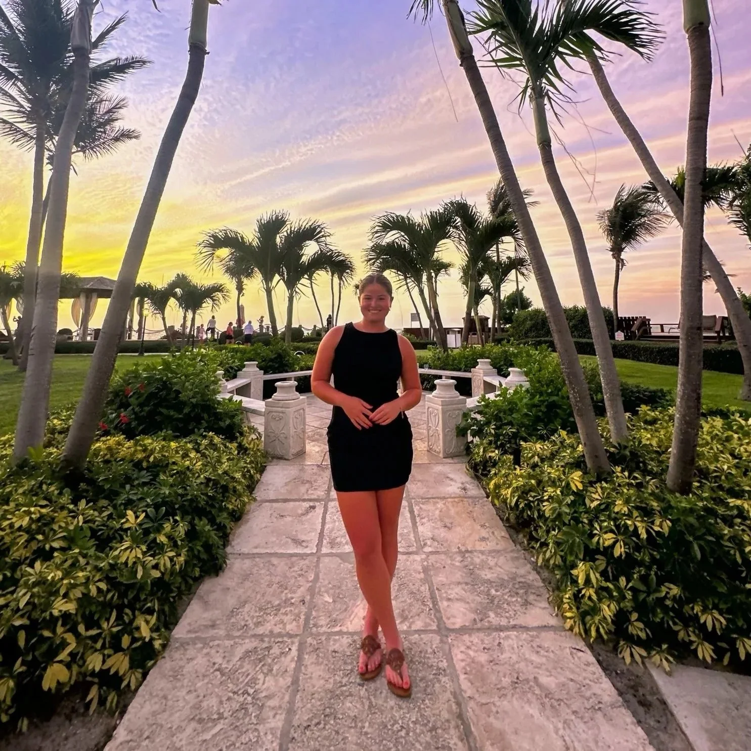 A woman in a black dress standing on a stone pathway in a tropical garden at sunset with palm trees and colorful sky.