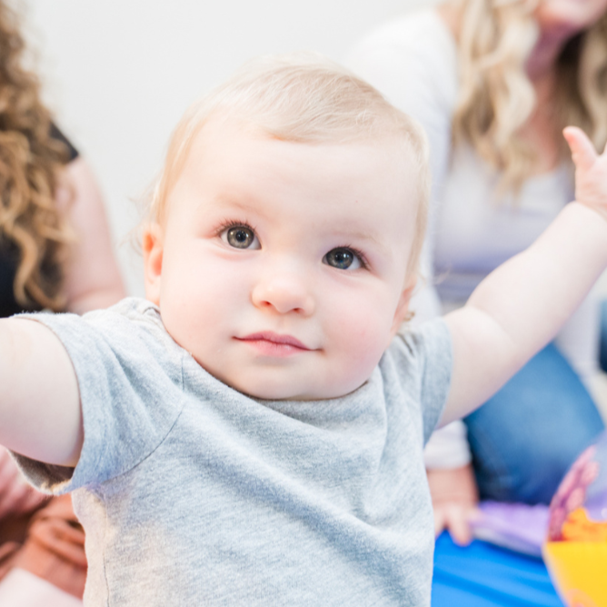 Close-up of a young child with blonde hair and blue eyes, wearing a gray shirt, extending arms outward.