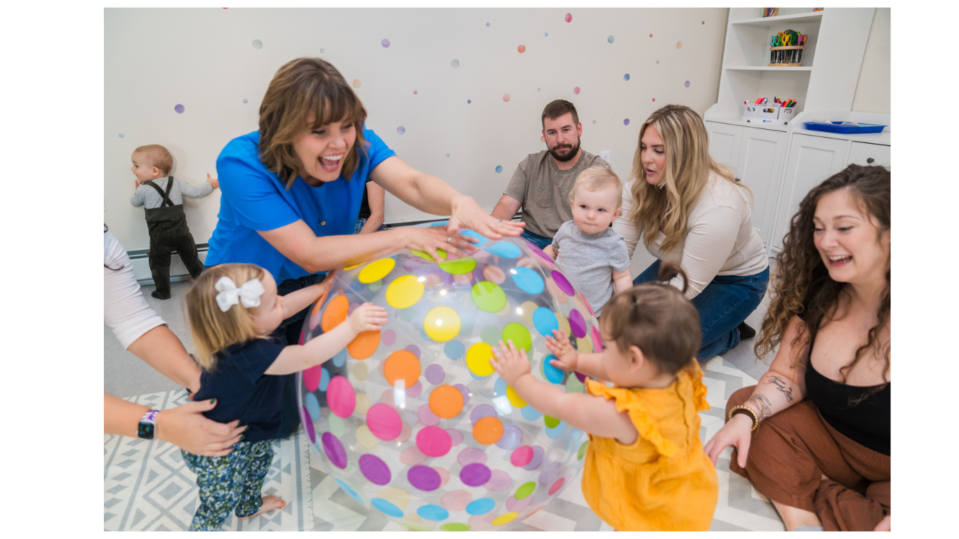 Group of children and adults playing with a large, colorful polka-dotted beach ball in a playroom.