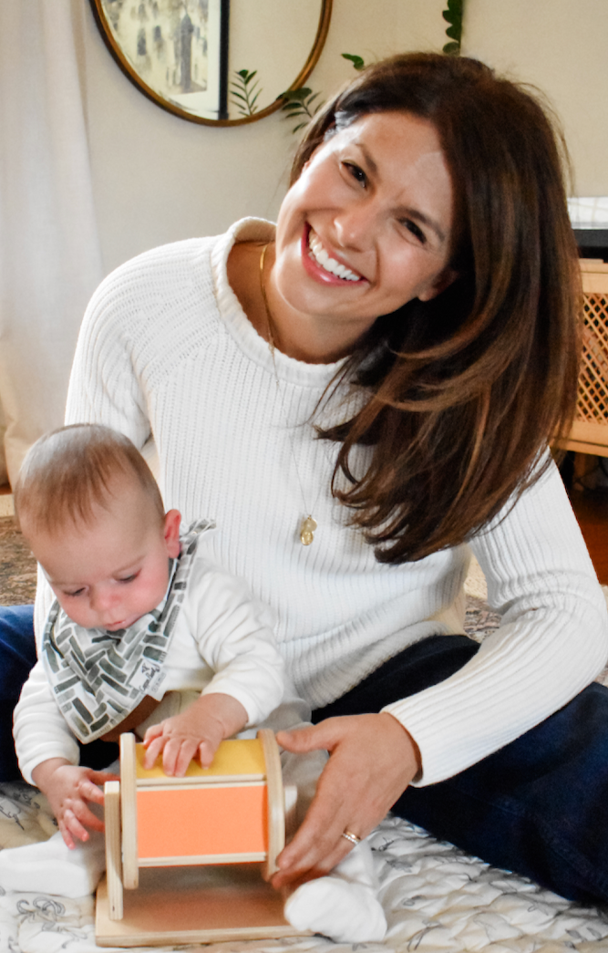A woman with long brown hair smiling and sitting on a bed with a baby boy. The baby is playing with a wooden toy and wearing a bib. The woman is wearing a white sweater and a pendant necklace. There are mirrors and plants in the background.