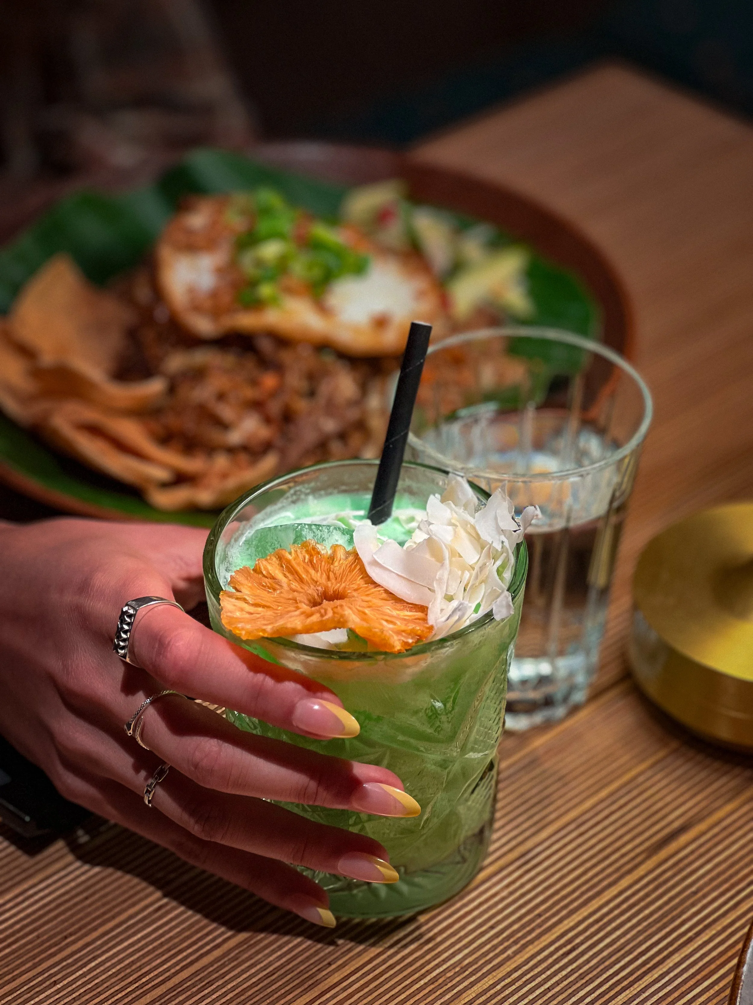 Een hand met goudgele nagels en rings houdt een groene cocktail met gedroogde sinaasappel en witte bloemen, op een houten tafel naast twee glazen en een gouden object, met een bord met vlees en garnituren op de achtergrond.