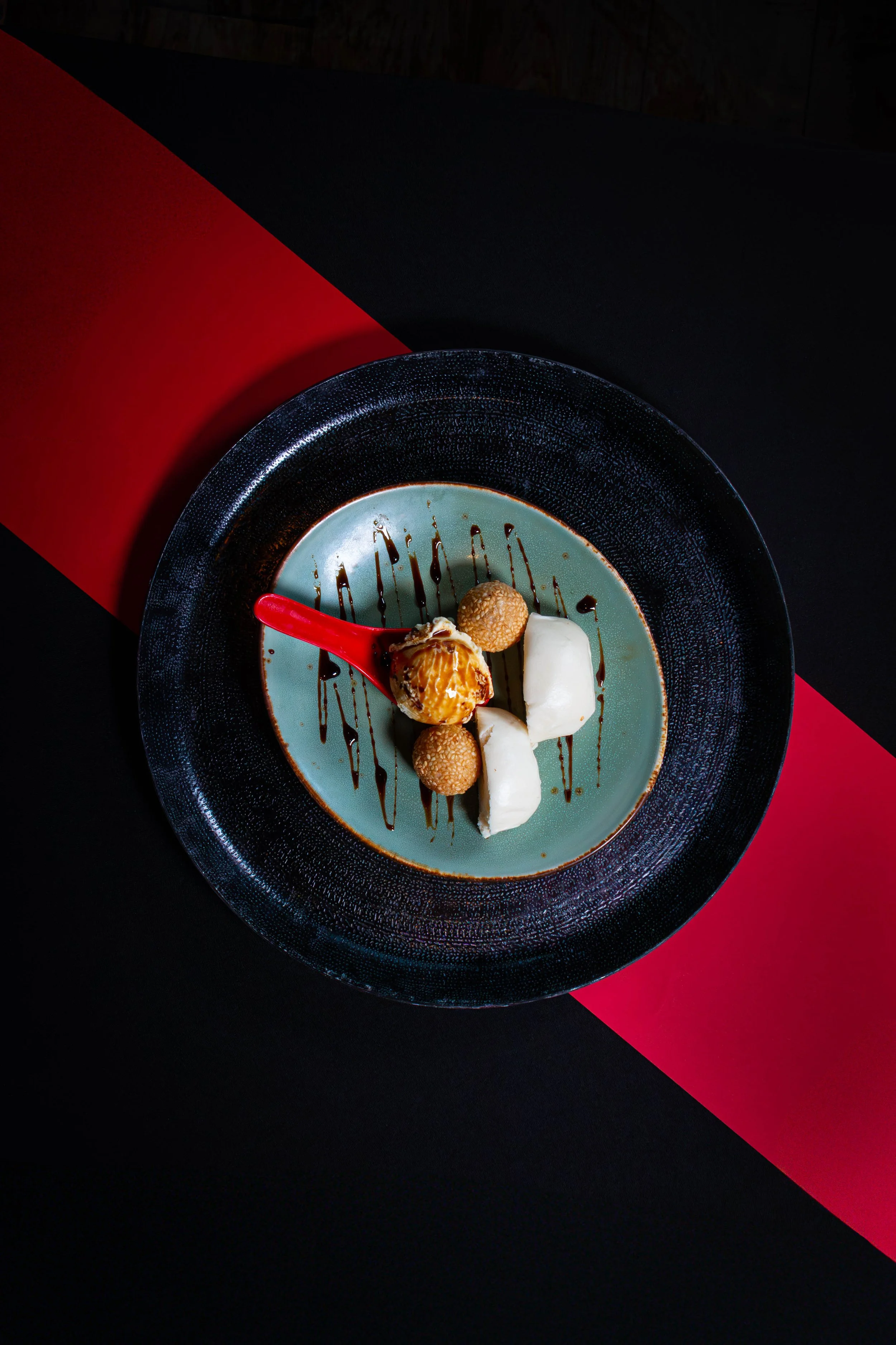 Gepresenteerde dessert met ijs, witte room, kleine koekjes, chocolade-siroop en een rode lepel op een blauwe en zwarte vlakke ondergrond.