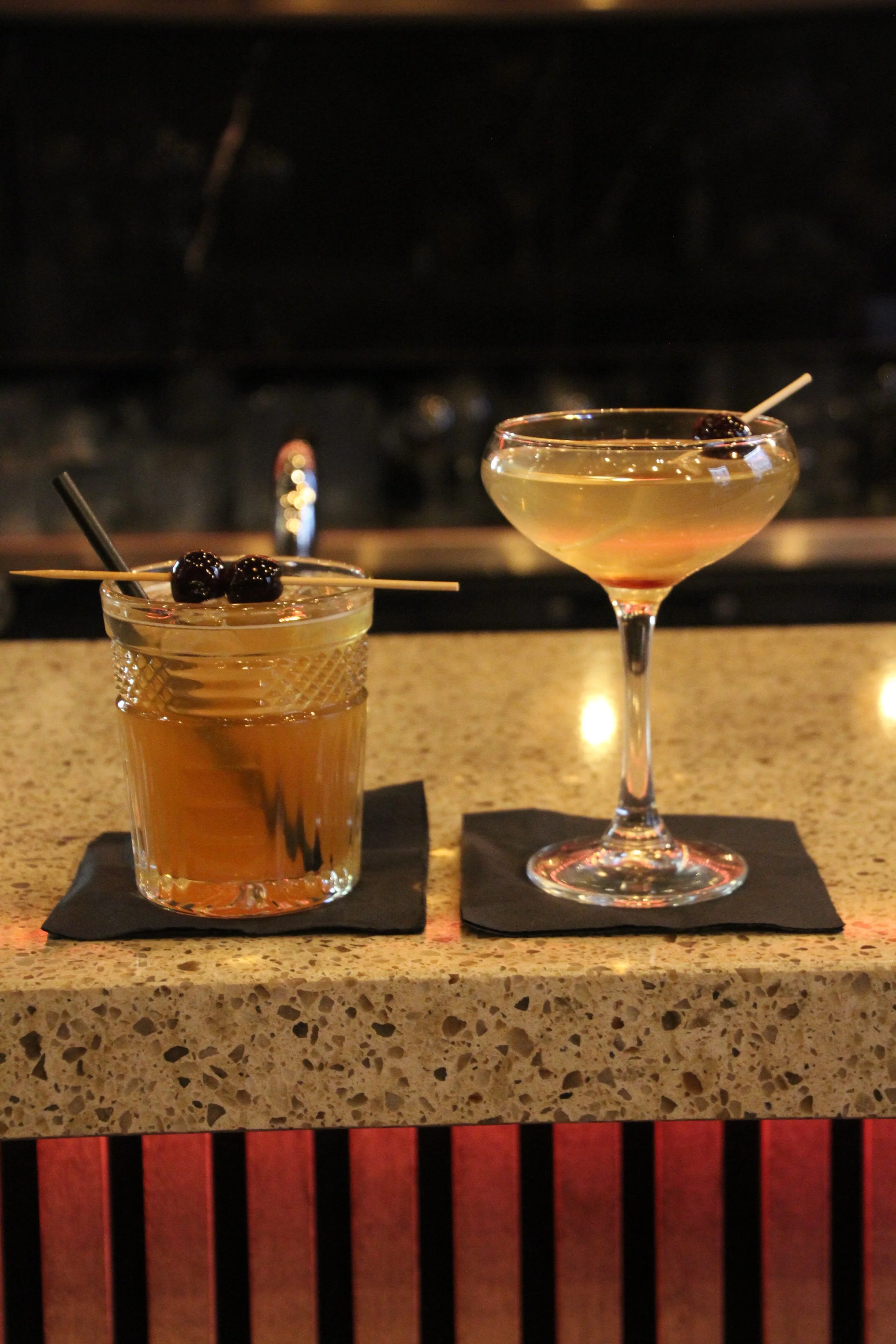 Twee cocktails, een whisky sour en een martini, op een bar counter met zwarte servetten eronder.