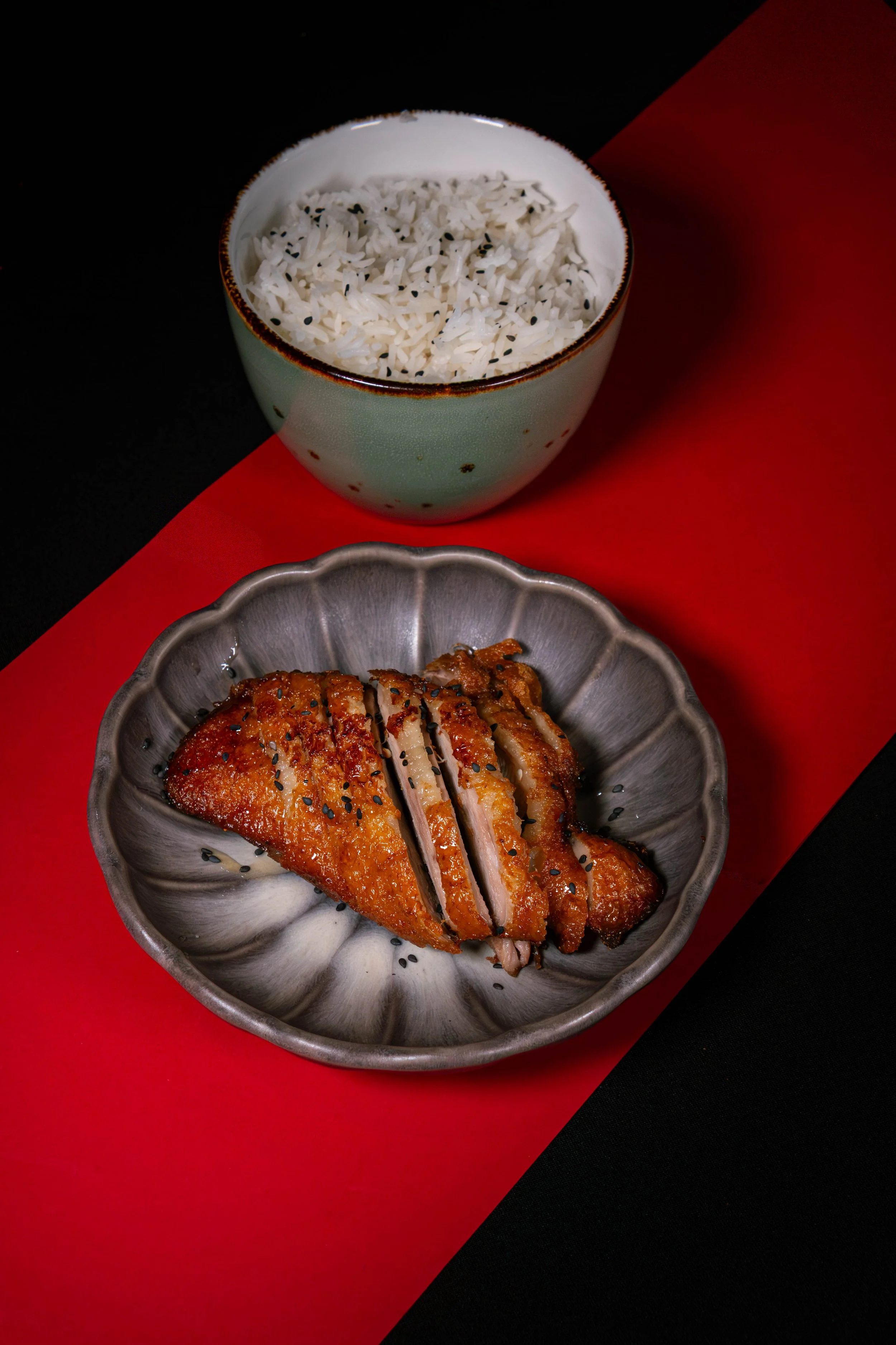 Gekookte rijst in een groene kom en knapperige eend op een grijs bord, beide op een rode en zwarte achtergrond.
