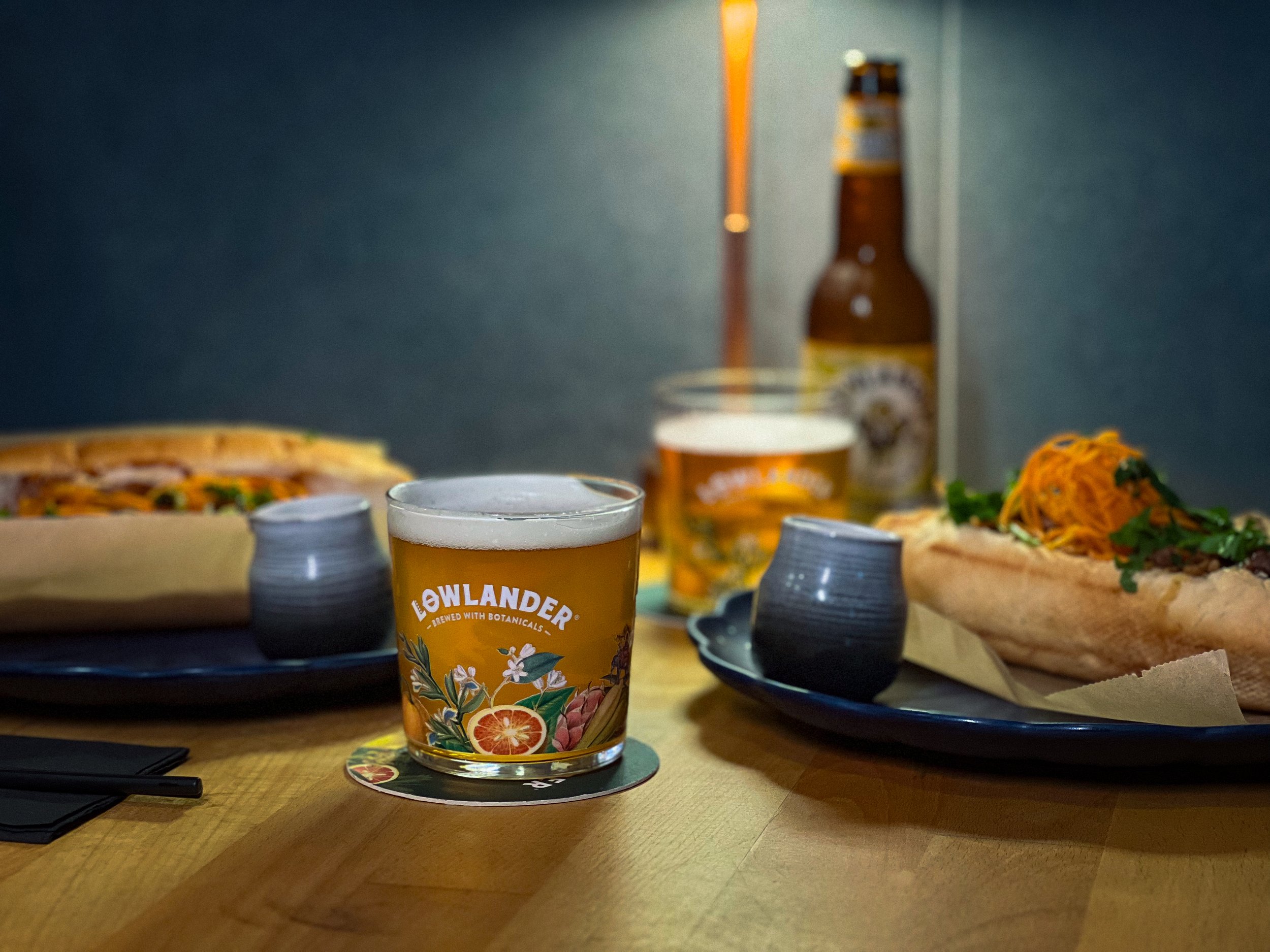 Twee glazen ambachtelijk gebrouwen bier met het logo 'LEWLANDER' op een houten tafel, omringd door verschillende gerechten zoals pizza en een broodje met geraspte wortel en groenten, met een donkere achtergrond en een lamp.