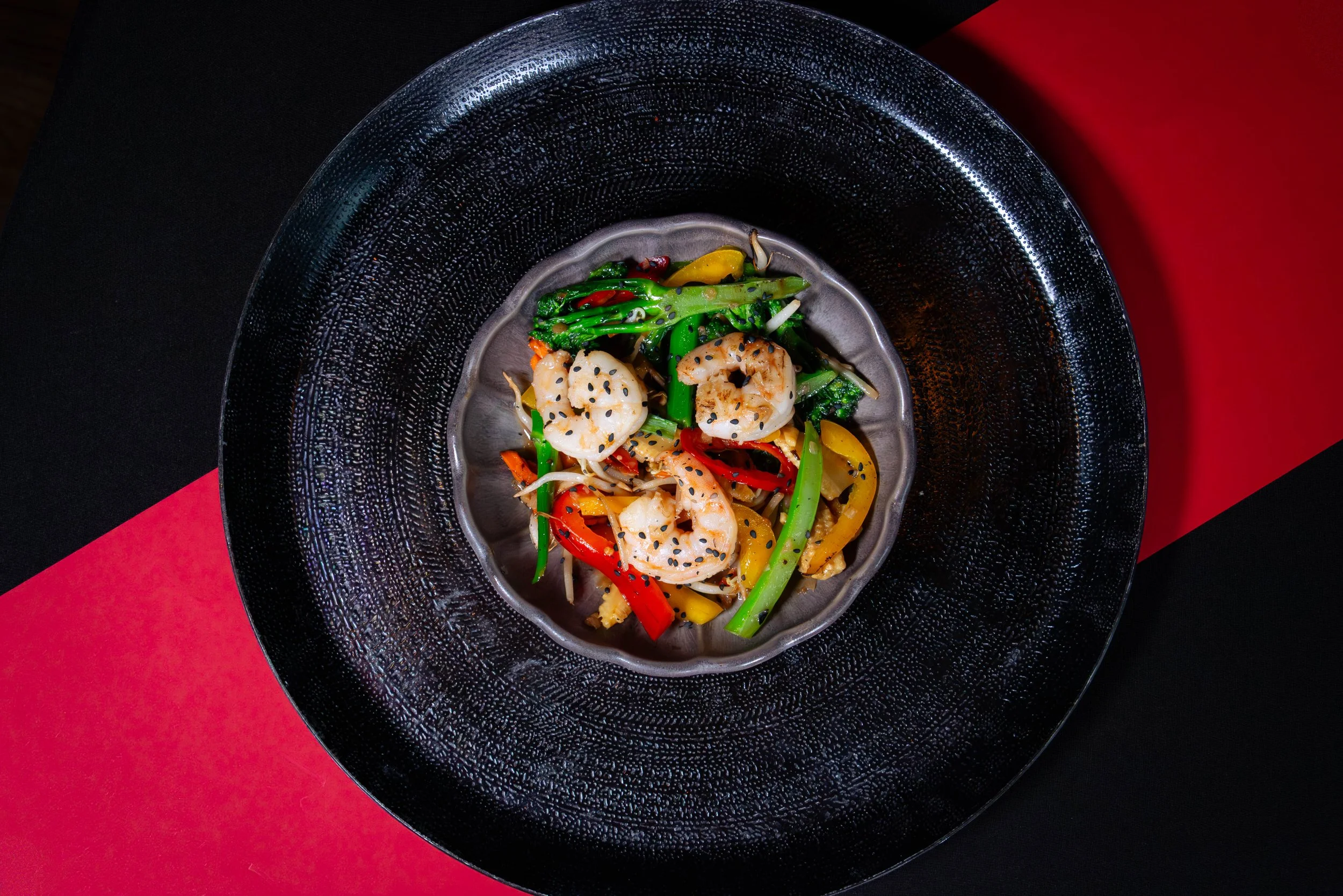 Gestoofd groente met garnalen en sesamzaad in een zwart bord op een zwart-rood achtergrond.