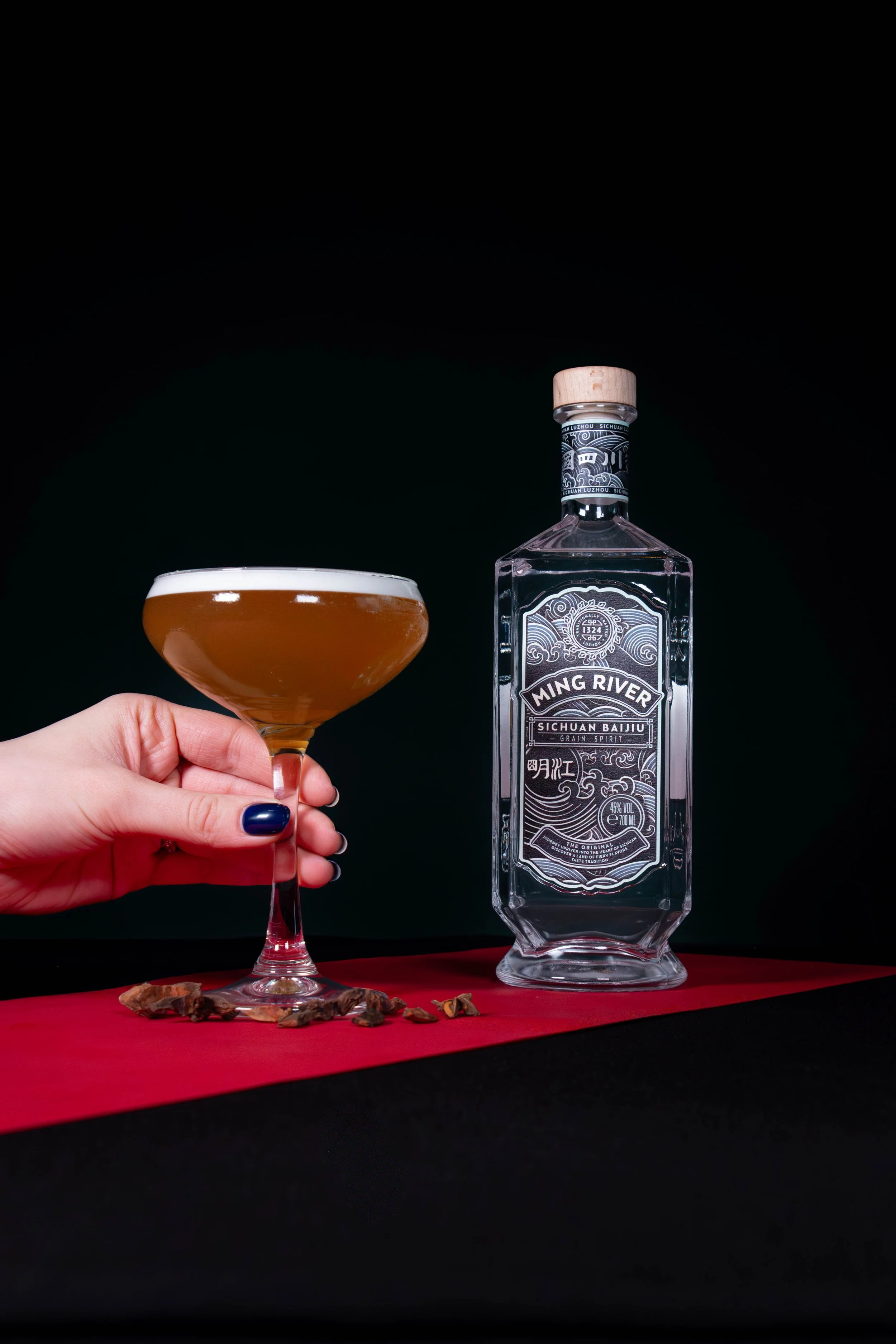 Een hand houdt een cocktailglas met een amberkleurige drank, naast een fles Ming River Sichuan Baijiu op een rode en zwarte ondergrond tegen een zwarte achtergrond.