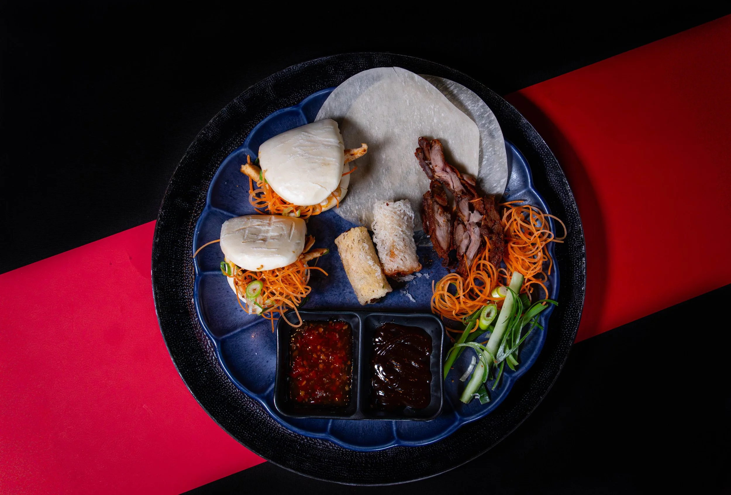 Een bord met gestoomde bao bun, plakjes geroosterde varkensvlees, gefrituurde varkenskoteletten, geraspte wortel, komkommer, groene uien, en twee kommen met saus, op een zwarte en rode achtergrond.