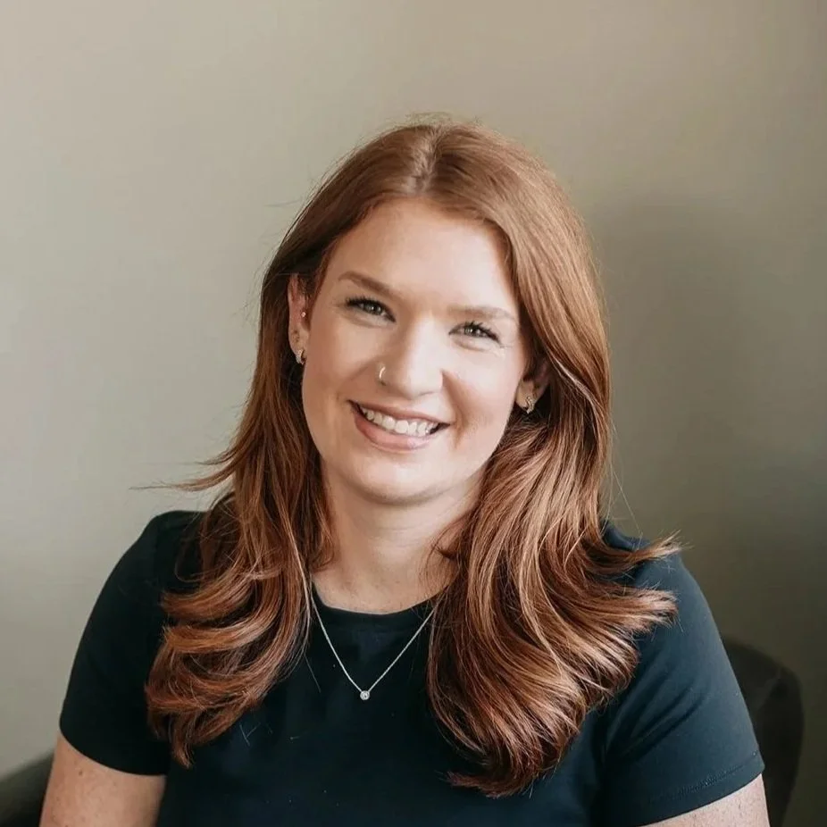 A woman with red hair smiling indoors, with a blurred background including a plant.