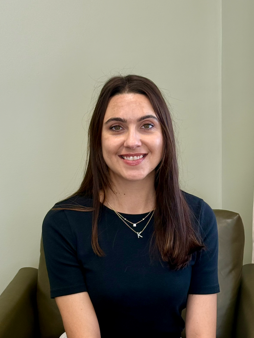 A woman with long brown hair and a black shirt smiling while sitting on a beige couch in a room with a neutral wall.