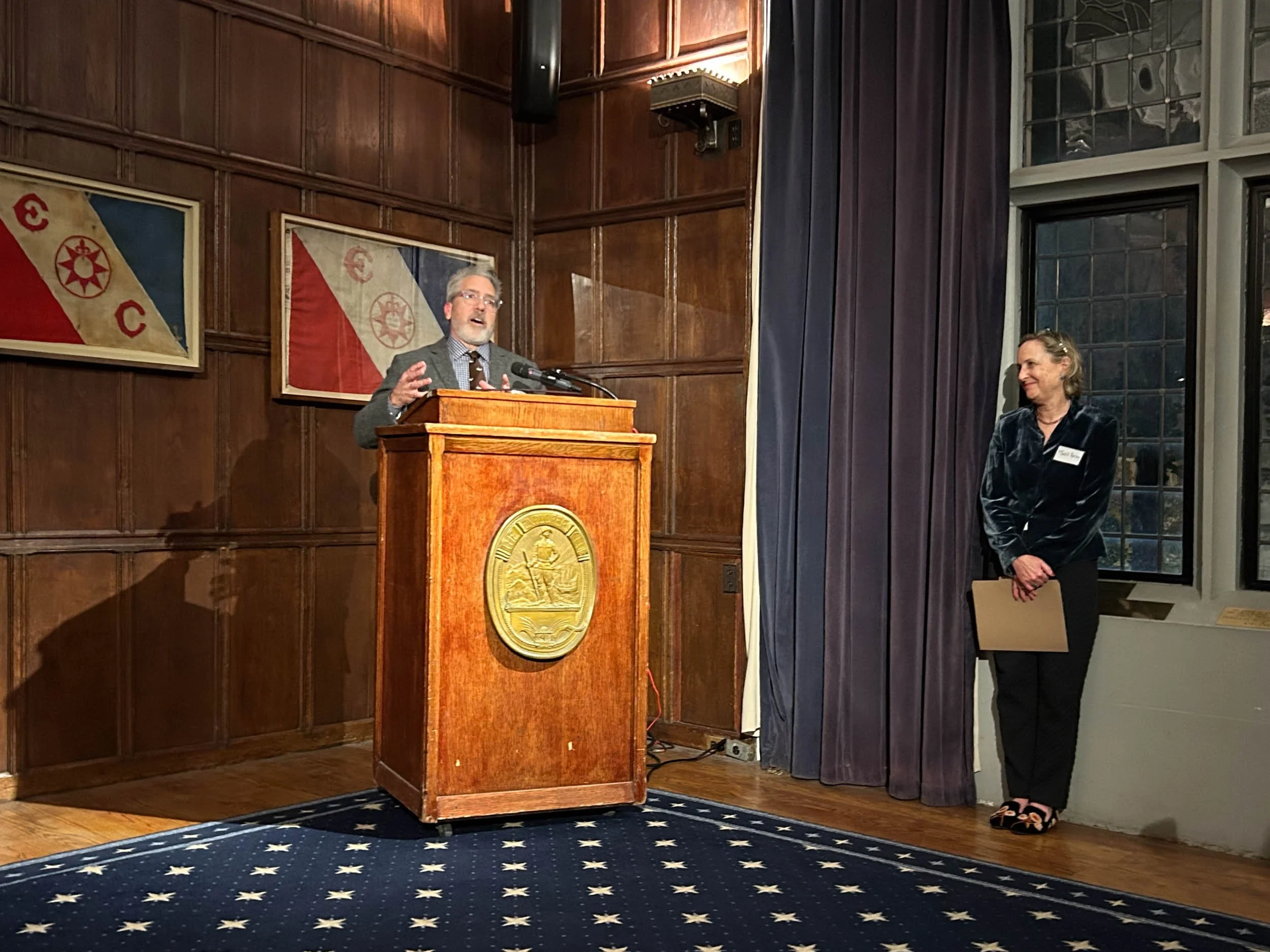 A man speaks behind a podium in a large, wood-paneled room