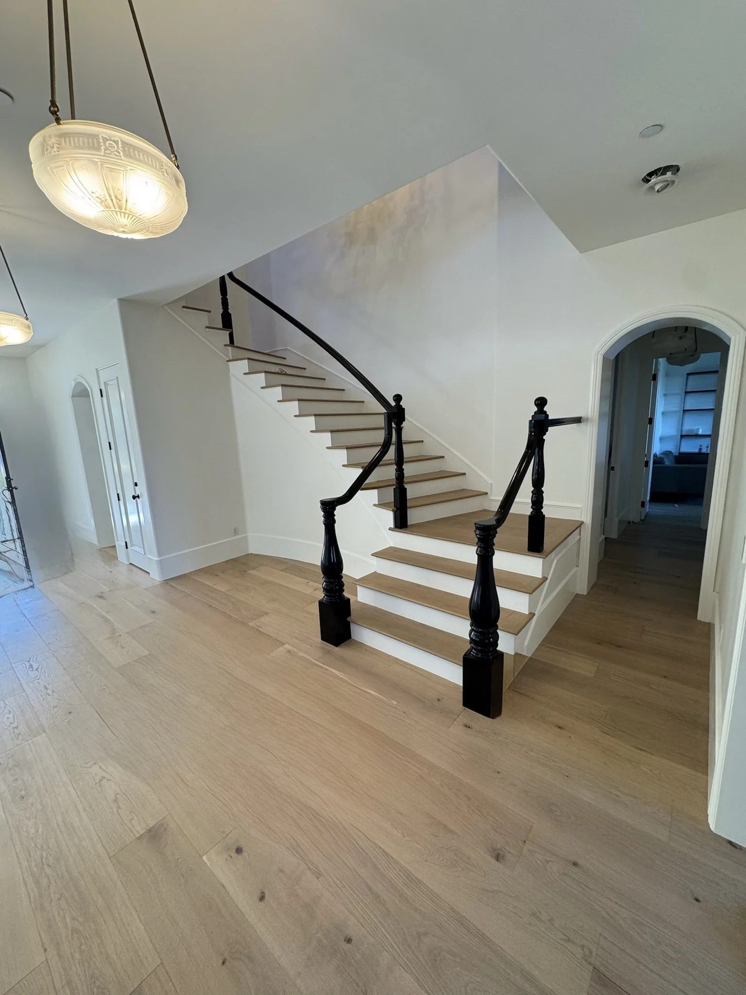 Interior view of a home with a wooden staircase featuring black handrails, light wood floors, chandelier lighting, and a hallway leading into other rooms.