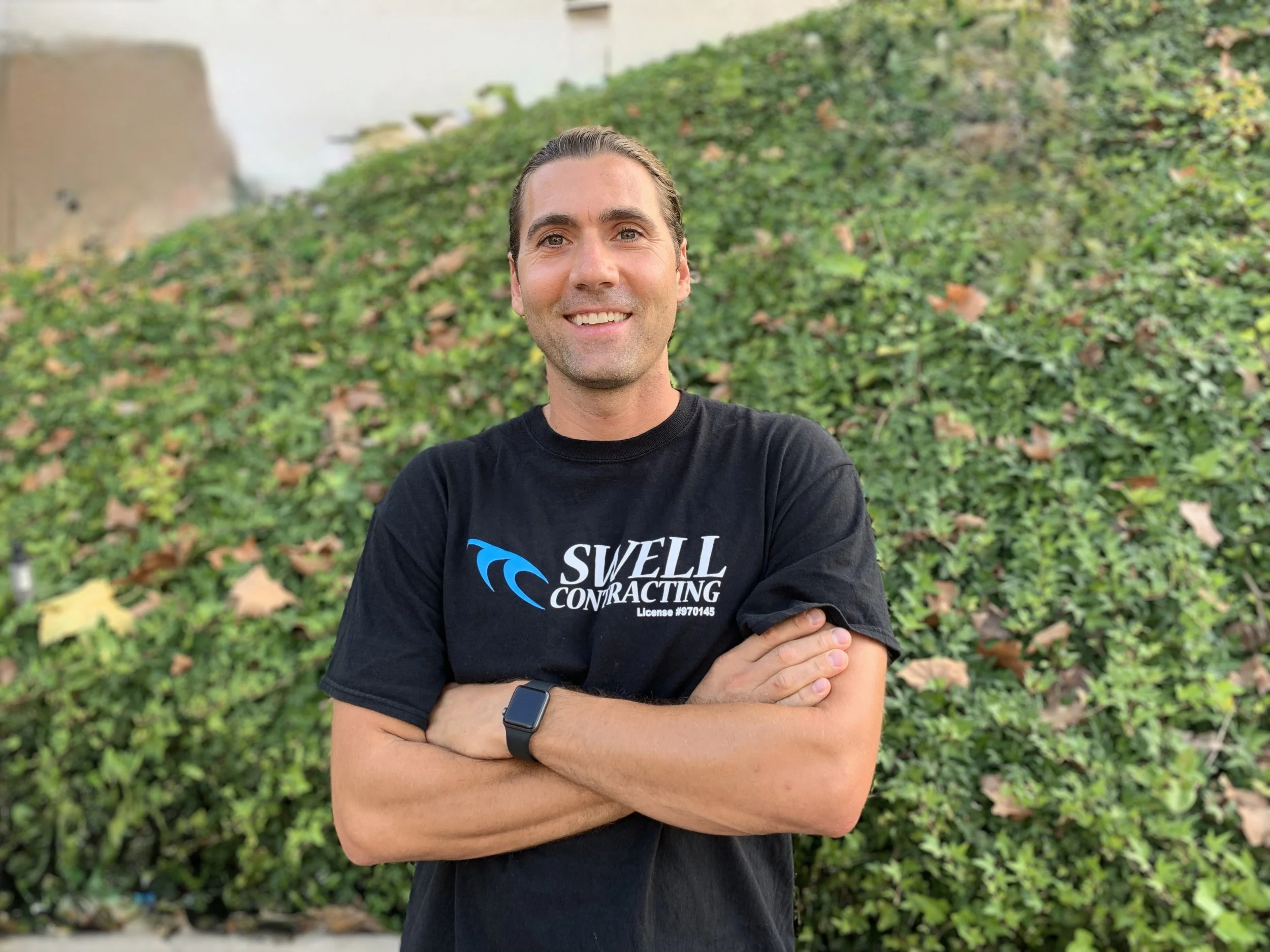A man with brown hair and blue eyes smiling, wearing a black T-shirt with 'Swell Contracting' logo, standing outdoors with crossed arms in front of green shrubbery.