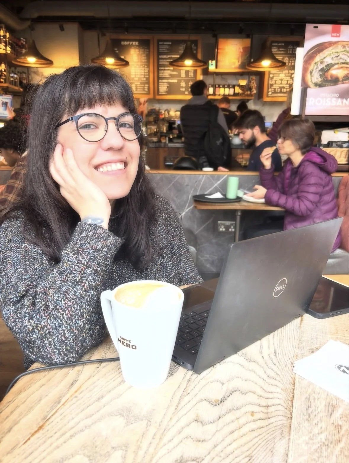 A woman with glasses smiling at the camera, sitting at a wooden table in a coffee shop with a cup of coffee, a Dell laptop, and a smartphone in front of her. The background shows the coffee shop counter with menu boards and other customers.