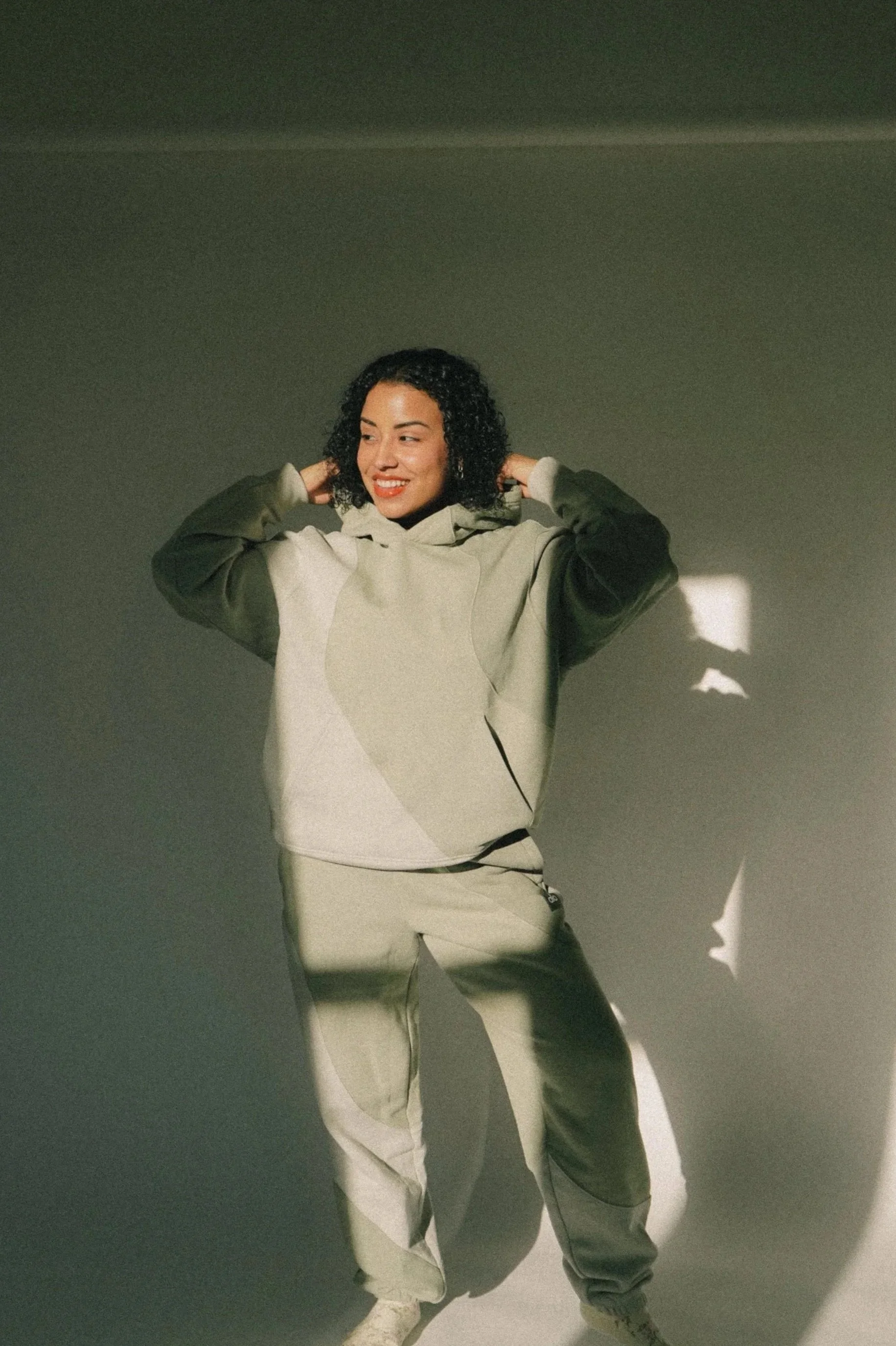 A woman with curly black hair wearing a light-colored hoodie and matching sweatpants, smiling and standing against a plain wall with light and shadow patterns.