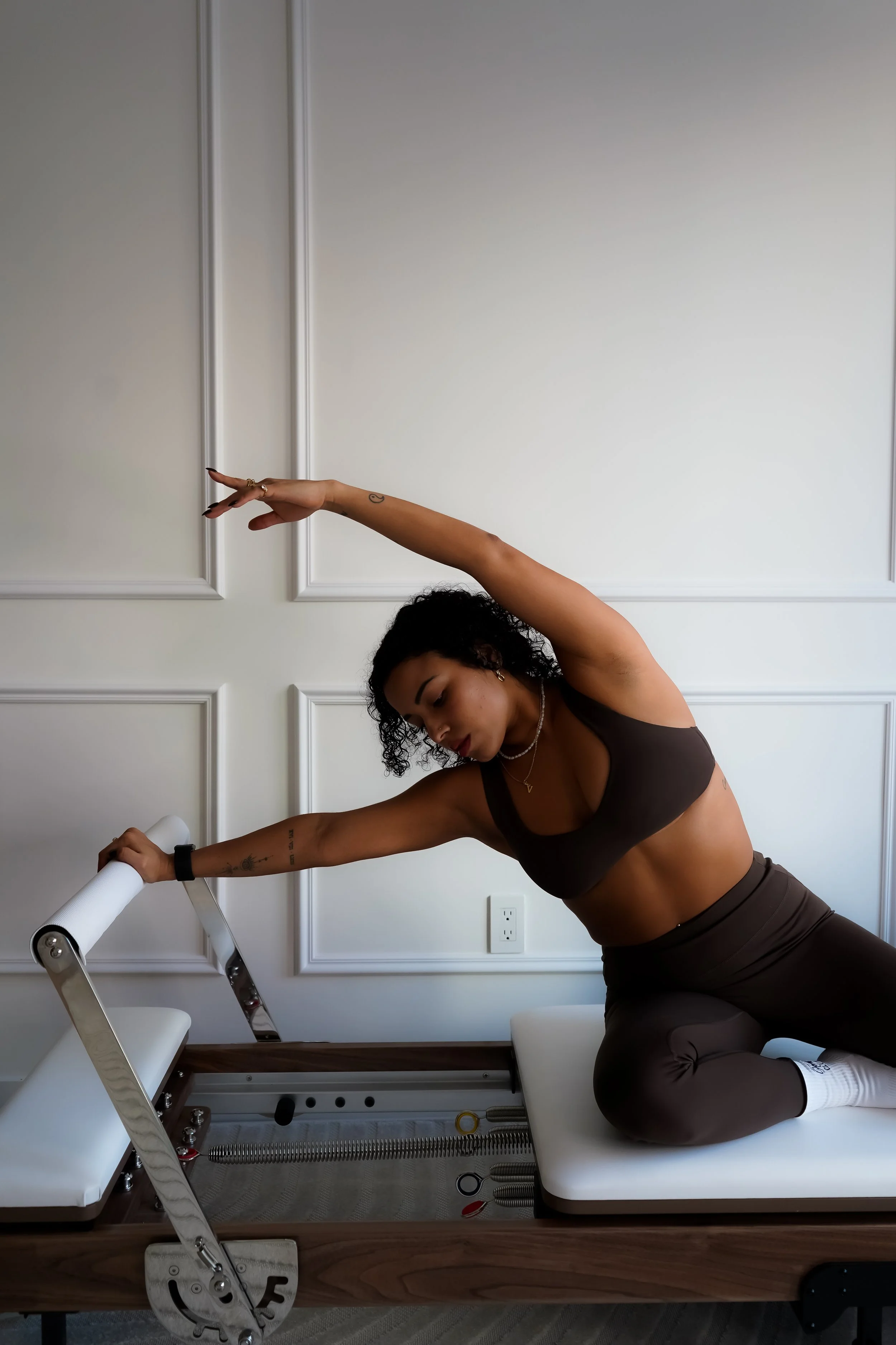 Woman in black sports bra and leggings performing a stretch on a Pilates reformer machine in a well-lit room.