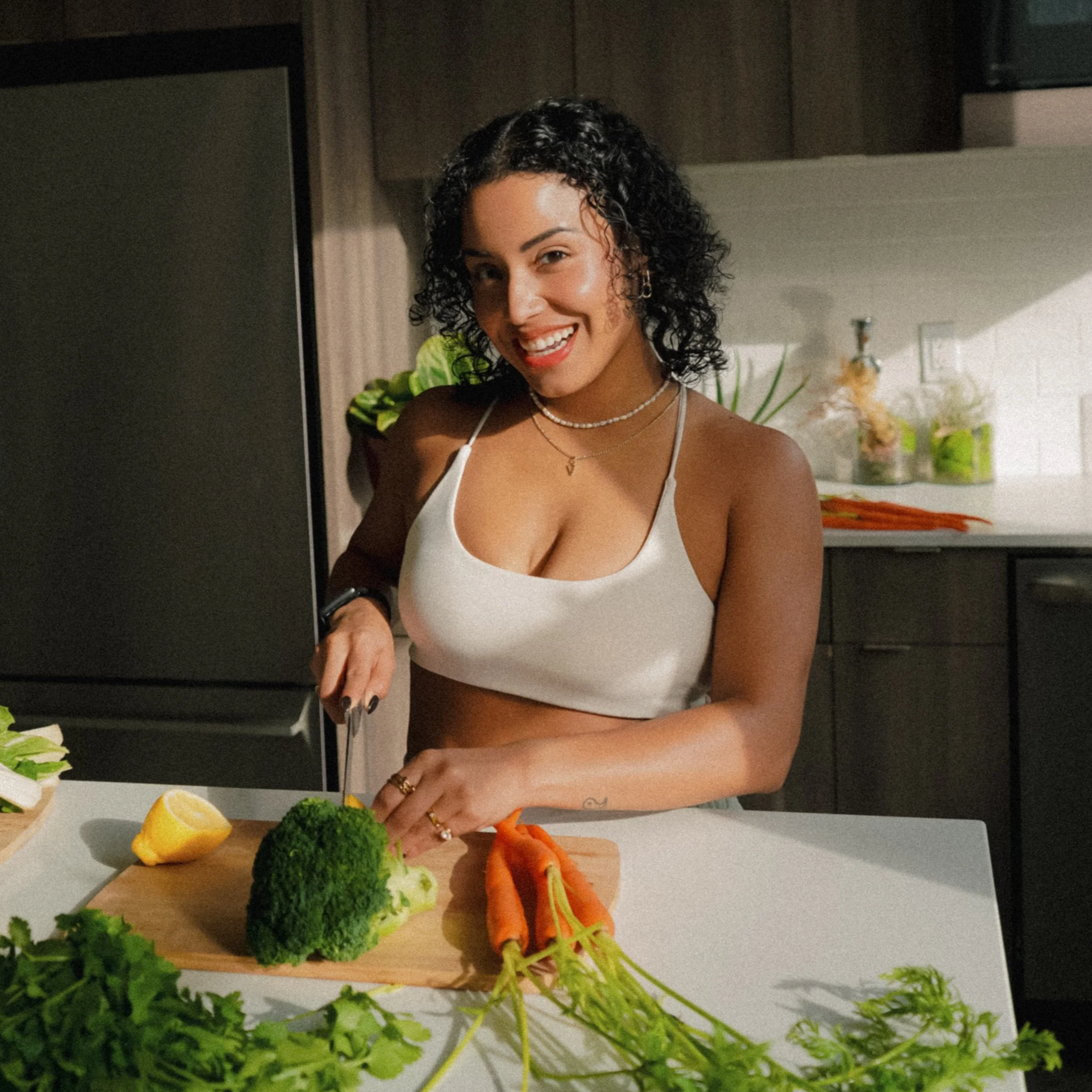 Woman with curly dark hair in a white crop top chopping broccoli and carrots in a modern kitchen.
