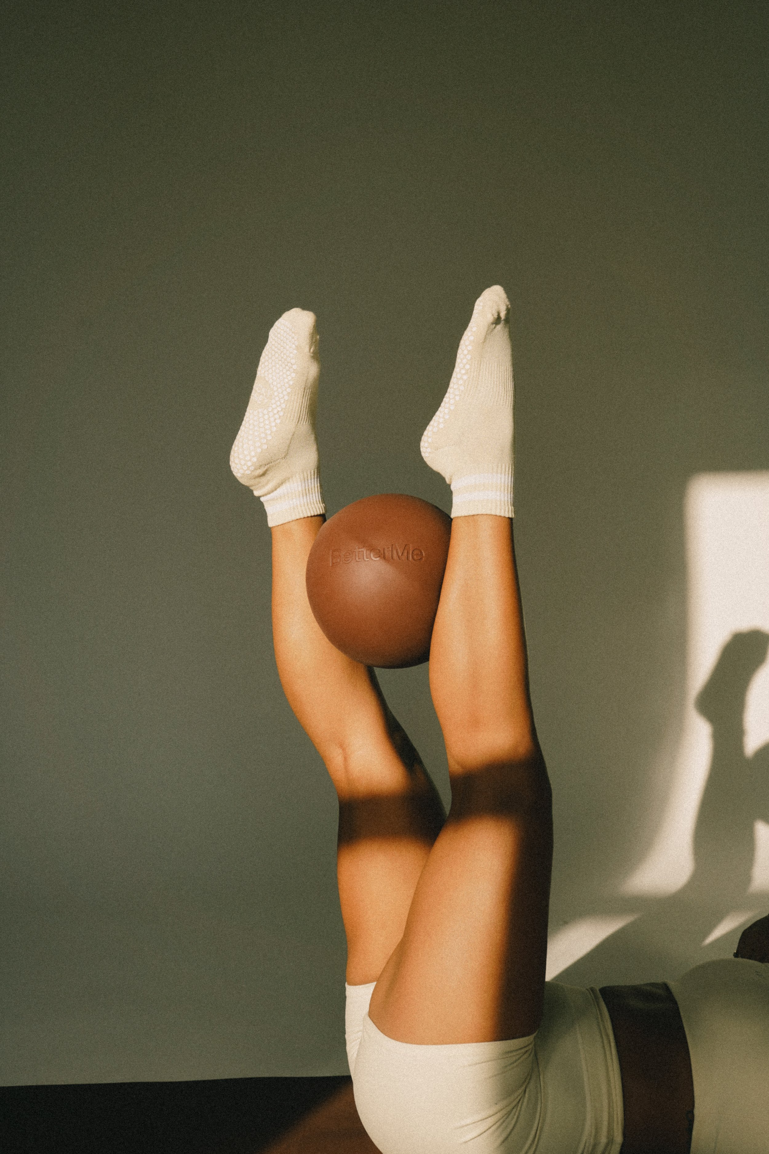 A person lying on their back with legs raised, holding a brown ball between their knees, wearing white socks and shorts, with sunlight casting shadows on the wall.