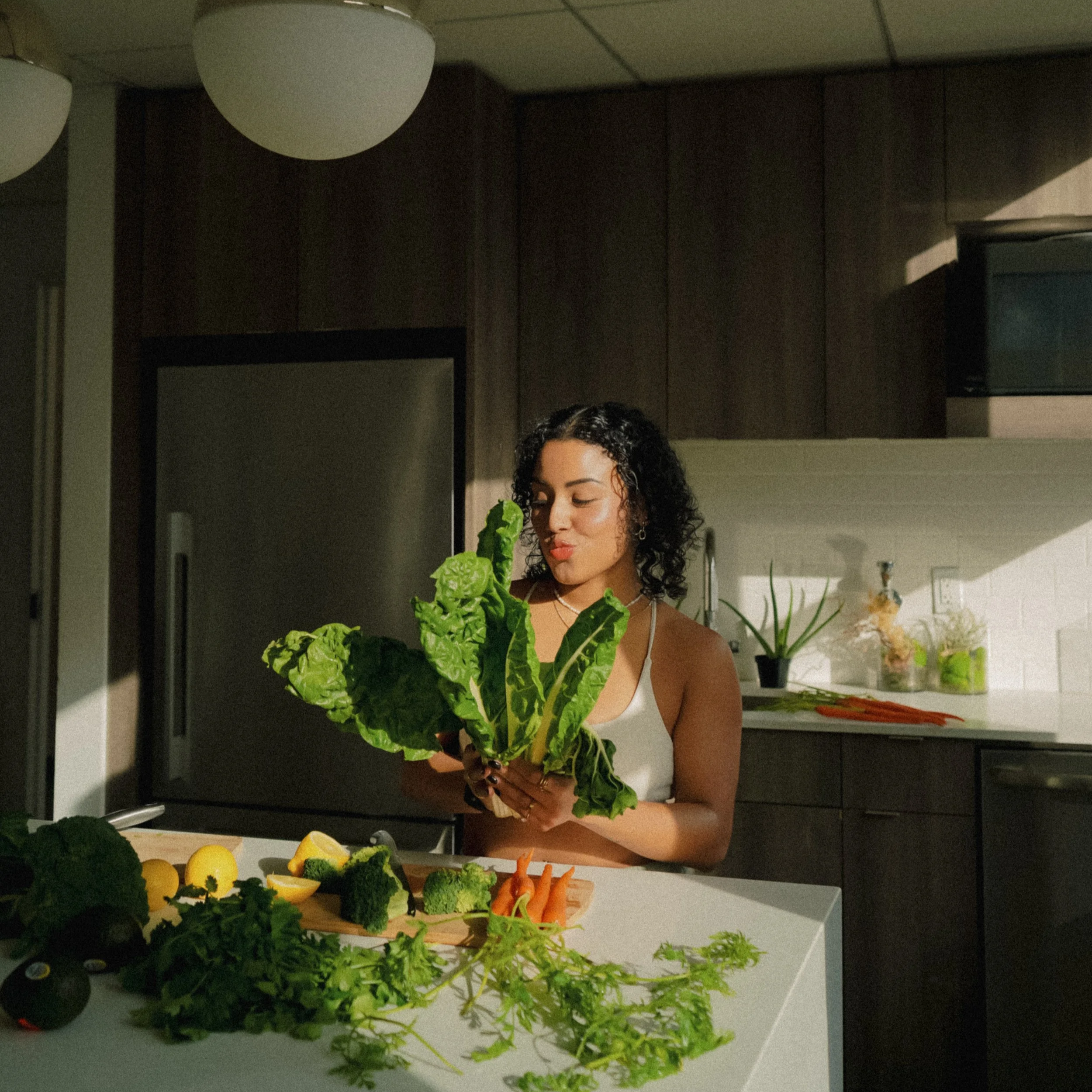 A woman with curly black hair holds a bunch of leafy greens in a kitchen. The kitchen has dark wood cabinets, white countertops, and various vegetables, including carrots, broccoli, lemons, and avocados, spread on the counter.Pilates for high achieve