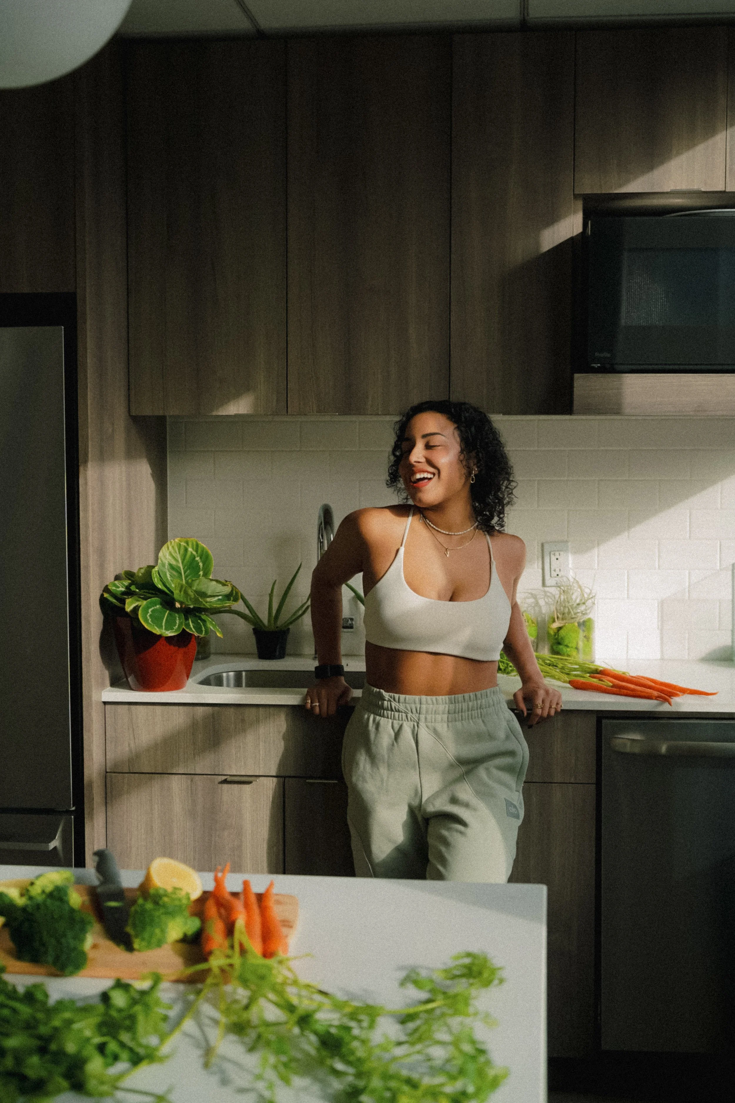 A woman with curly hair laughing, leaning against a kitchen counter surrounded by vegetables and plants.