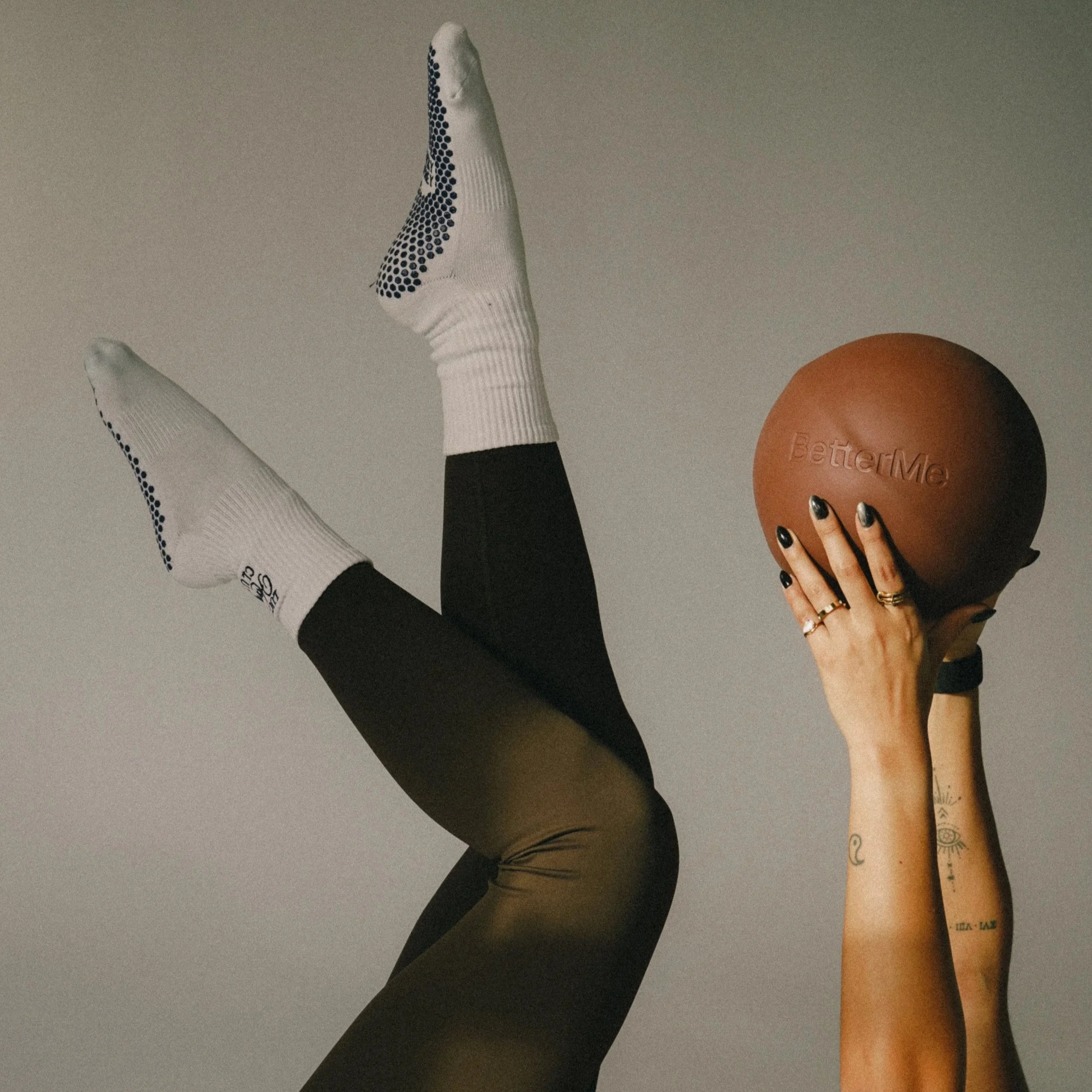 A person holding an orange exercise ball labeled 'BetterMe' with tattooed arms and black painted nails, legs raised wearing white athletic socks and black leggings.