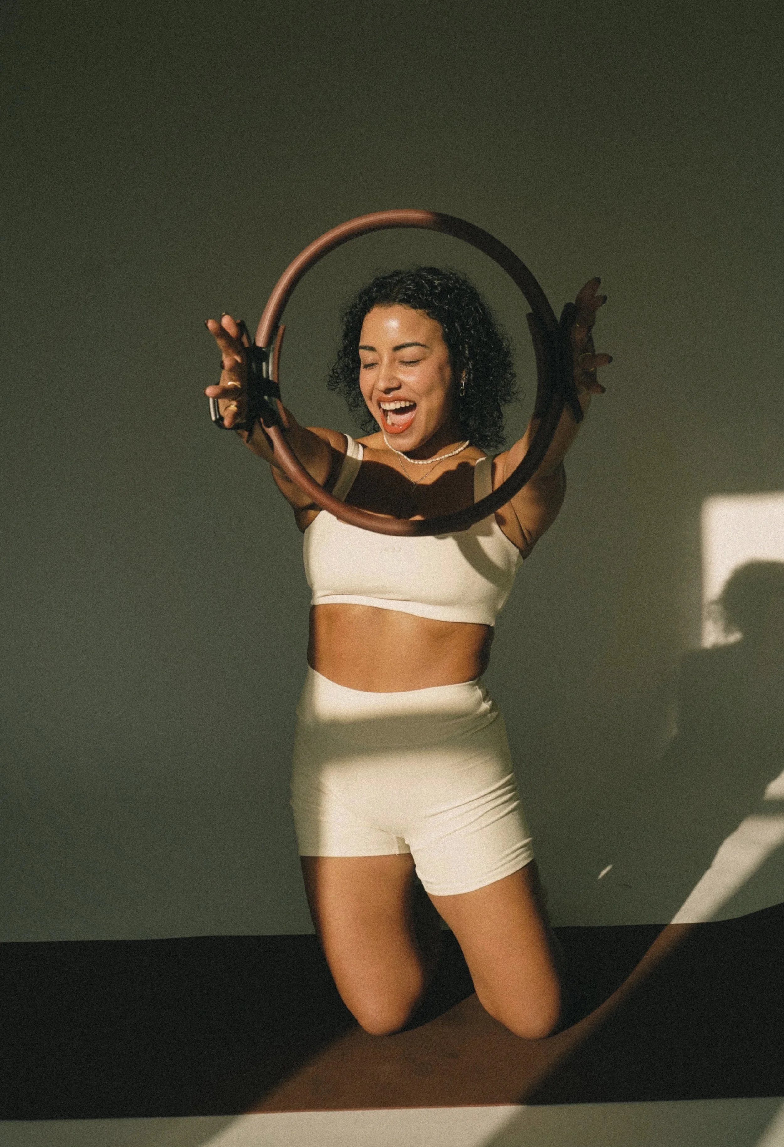 A woman with curly black hair, wearing a beige and black sports bra and matching shorts, is kneeling on the floor and holding a brown hula hoop with her eyes closed and a big smile.
