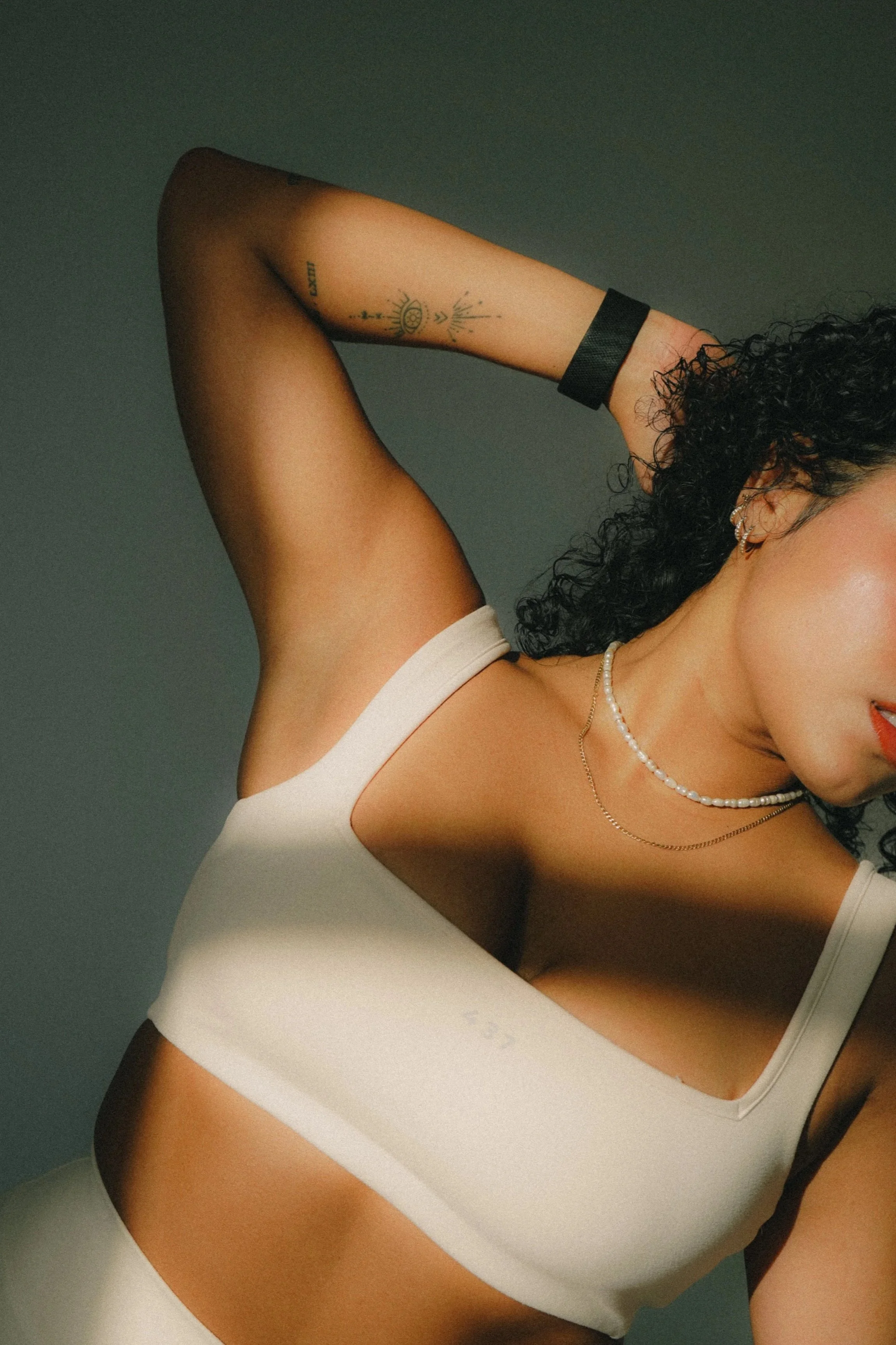 Close-up of a woman with curly hair, wearing jewelry, a white top, and a black wristband, against a dark background.