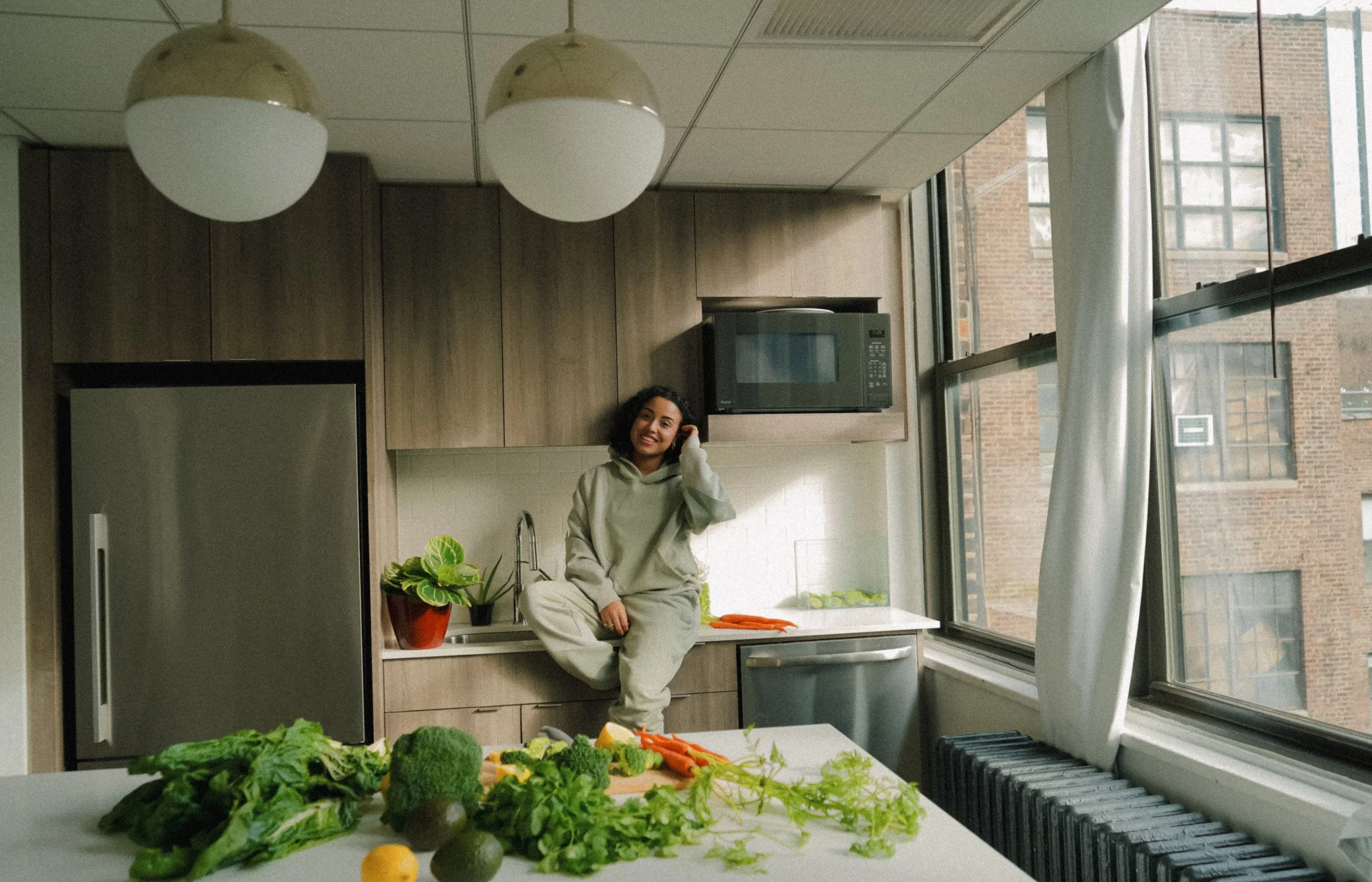 A woman sitting on a kitchen counter near a sink, surrounded by fresh vegetables and plants, in a modern kitchen with large windows and city views.