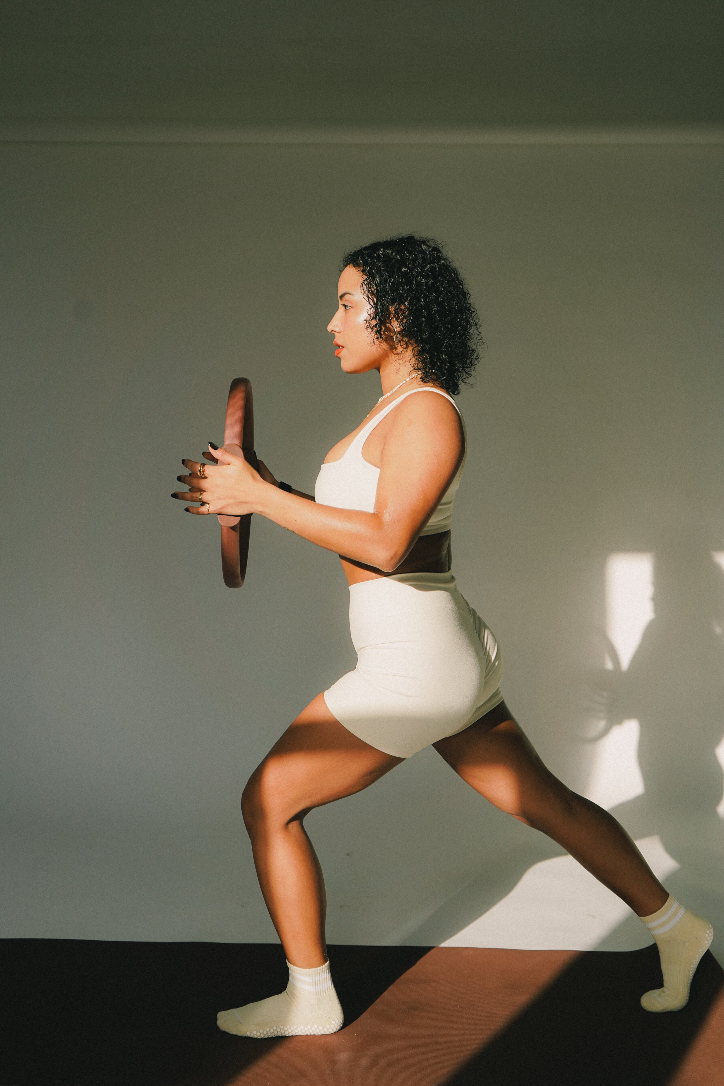 A woman with curly black hair in athletic clothing, holding a weight plate while performing a lunge. pilates instructor pose pilates photoshoot