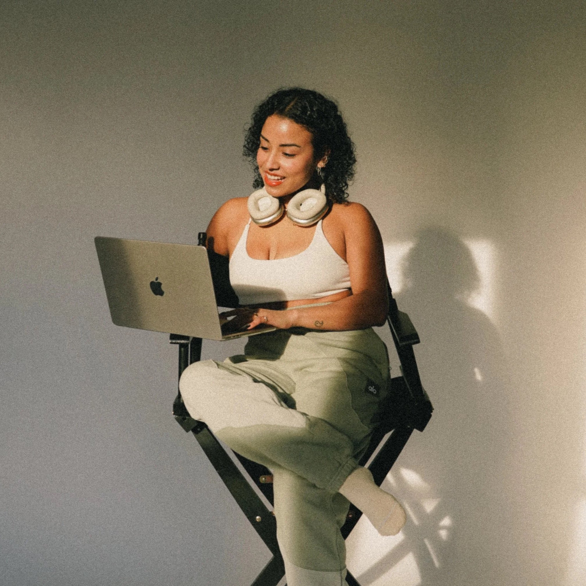 A young woman with curly black hair sitting on a director's chair, wearing a white tank top with headphones around her neck, working on a silver MacBook with a plain background.