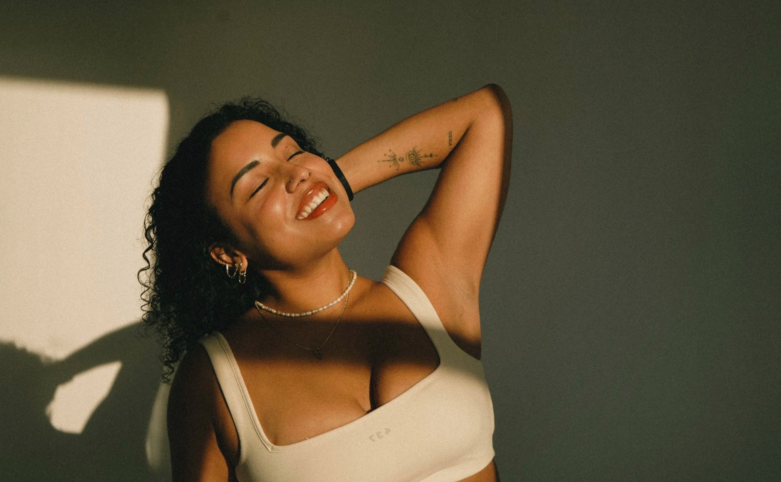 A young woman with curly hair posing with her eyes closed and a big smile, wearing a white tank top and jewelry, against a plain background with shadows.