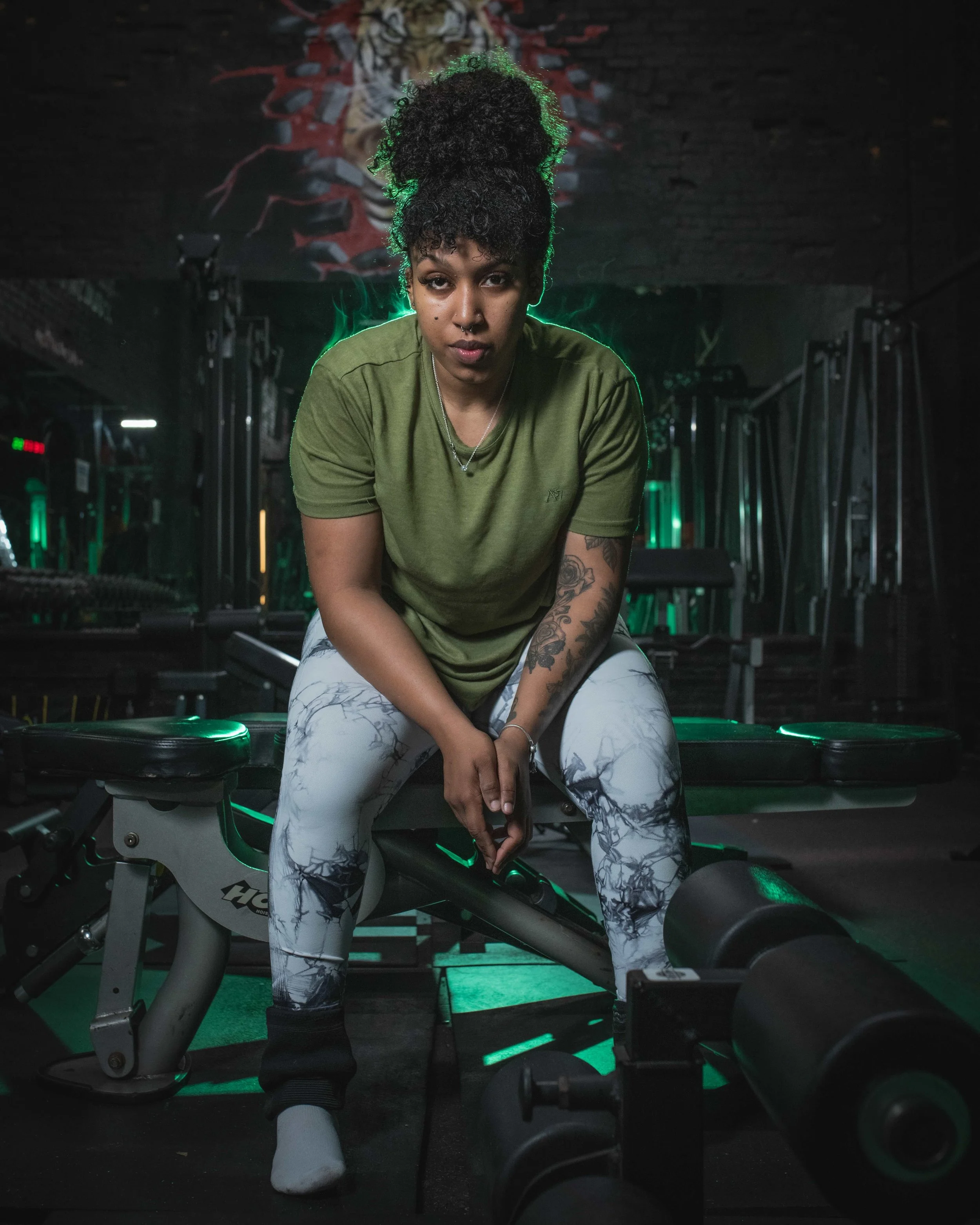 A woman in a green t-shirt and marble pattern leggings sitting on a workout bench in a gym with dark walls and green lighting.