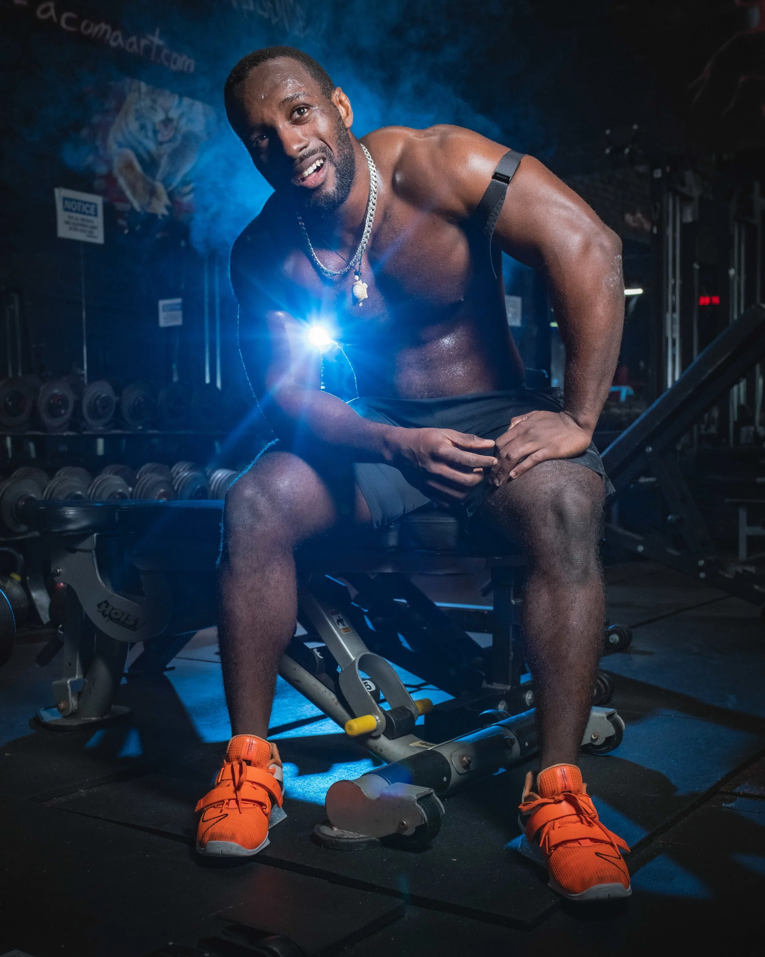 A shirtless man in workout clothes sitting on a bench in a gym, smiling at the camera, with weightlifting equipment in the background.