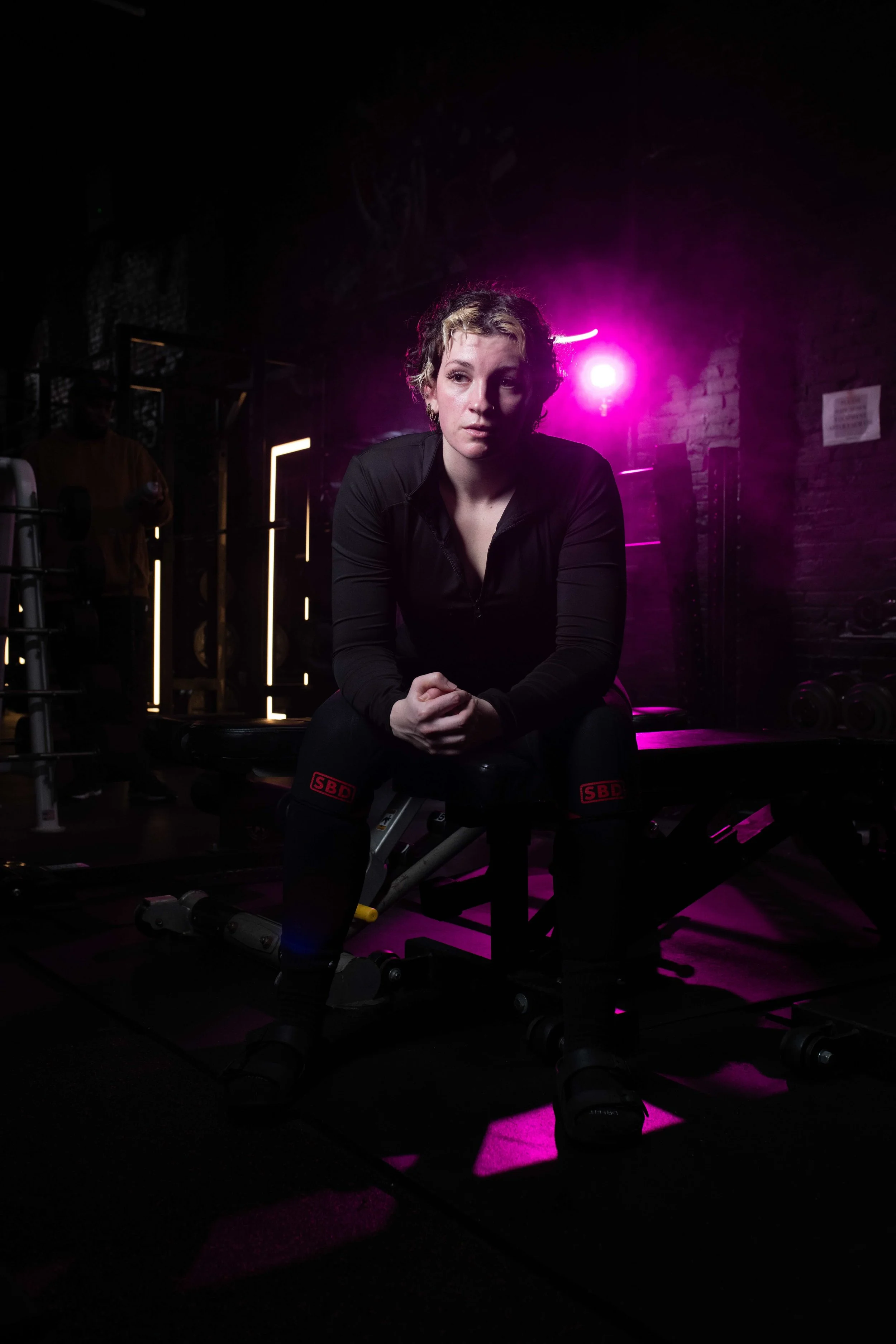 A woman with short blonde hair sits on a workout bench in a dimly lit gym with purple lighting, wearing black workout clothes.