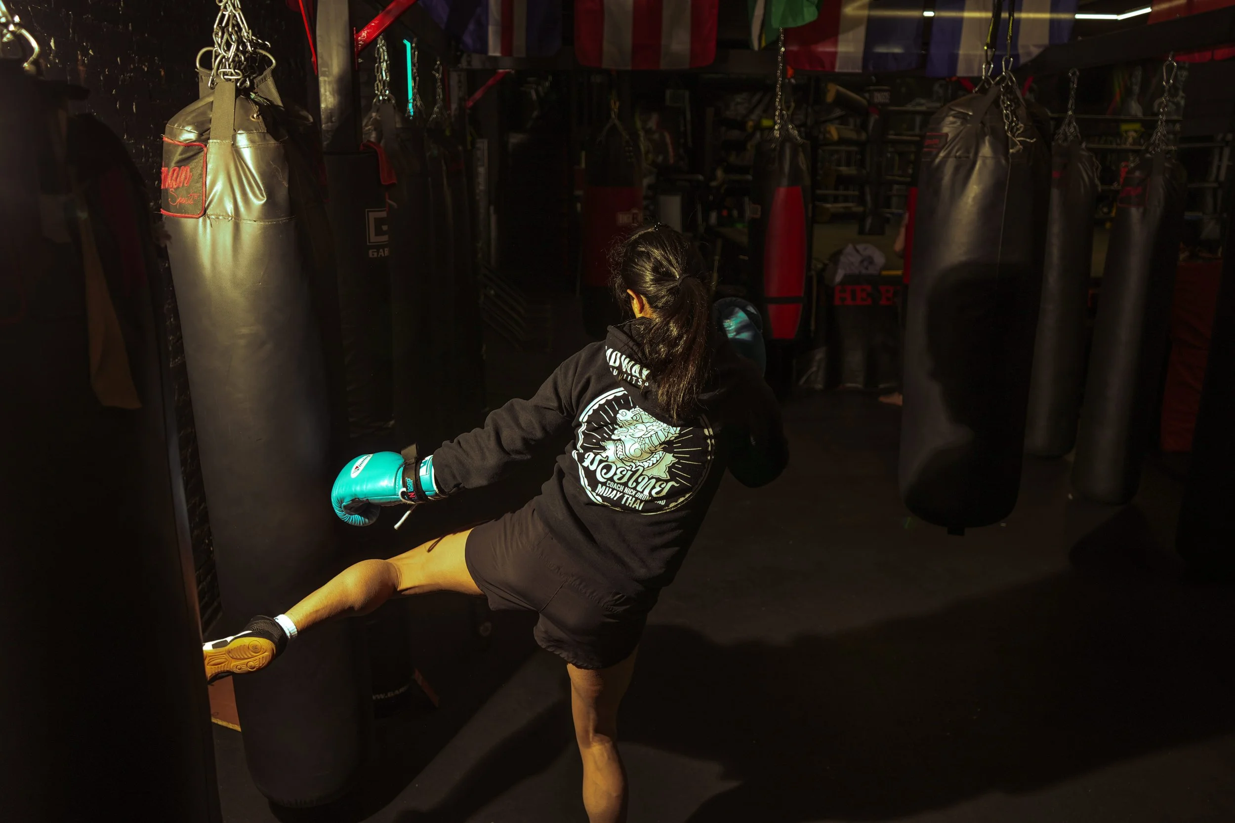 A woman practicing a kick on a punching bag in a boxing gym, wearing boxing gloves, a black hoodie, black shorts, and athletic shoes.