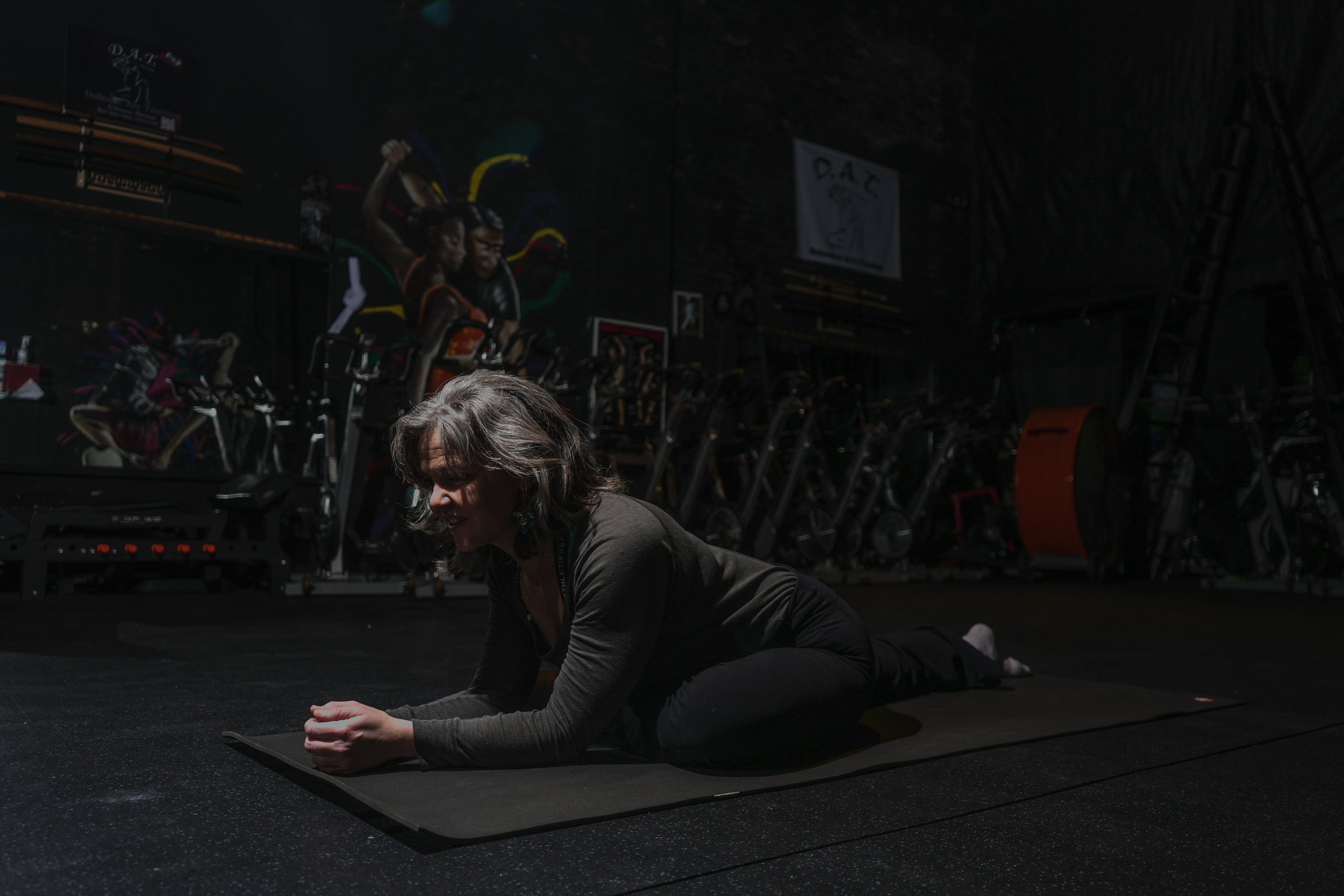 A woman practicing yoga on a mat in a dark gym, with exercise bikes and fitness equipment in the background.
