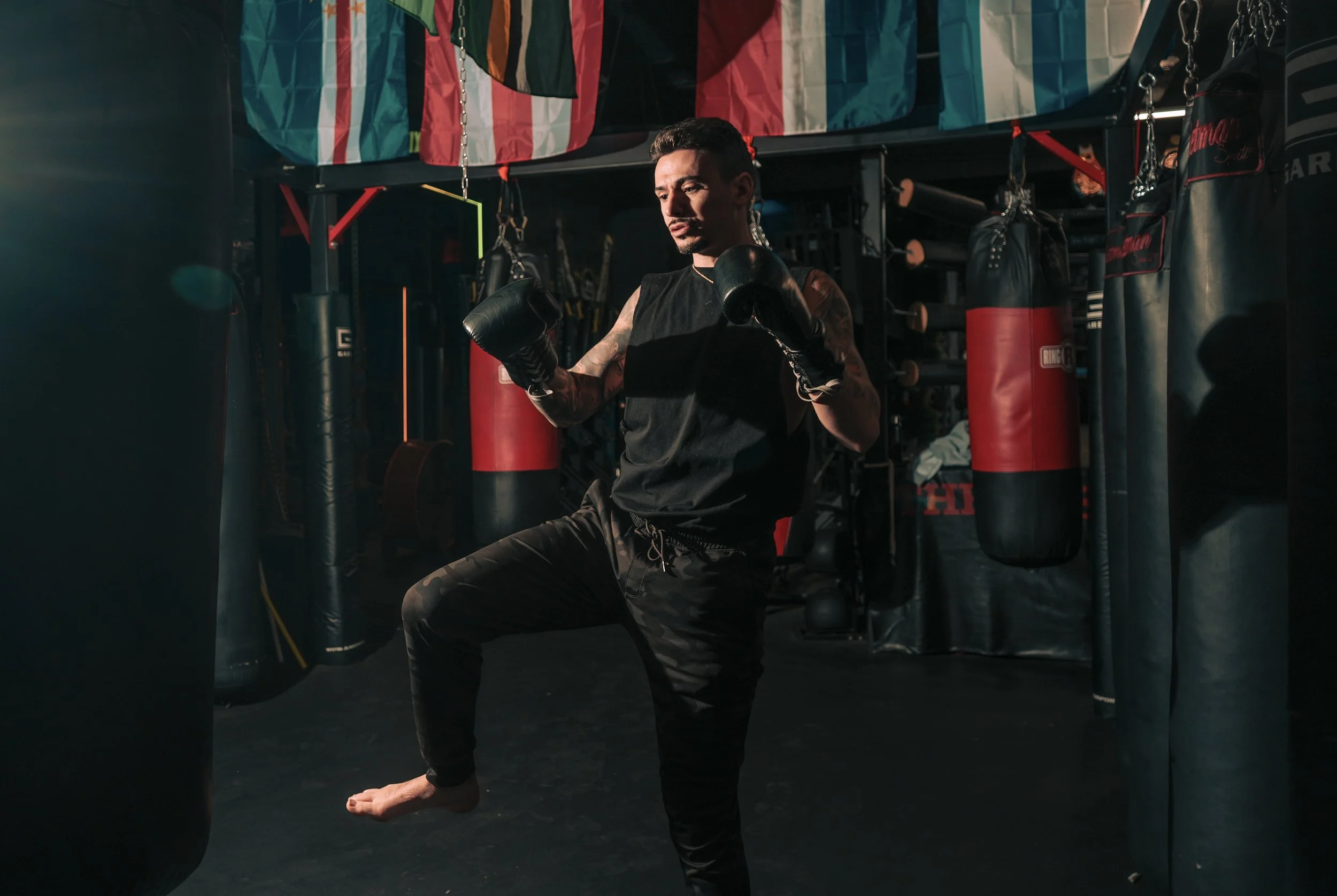 A man practicing boxing in a gym, wearing black gloves and workout clothes, surrounded by punching bags and gym equipment.