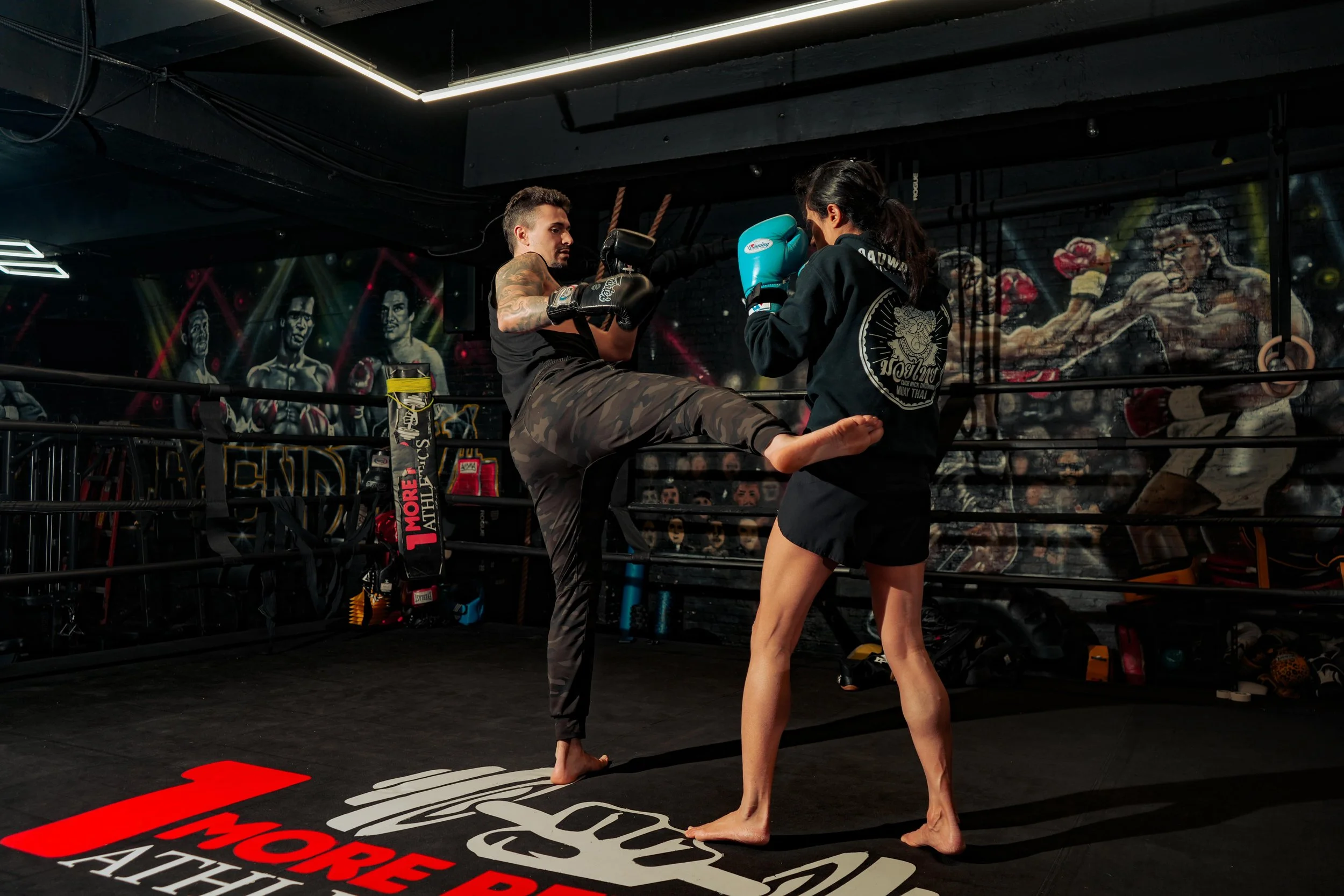 Two women practicing martial arts in a boxing gym, one executing a high kick towards the other, who is holding blue boxing gloves, with a mural of fighters on the wall in the background.