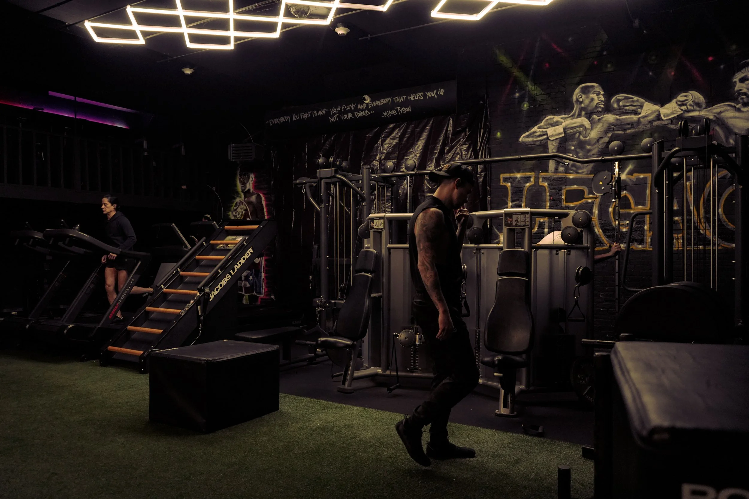 People working out in a dimly lit gym with wall murals of boxers and motivational quotes, including a woman on treadmill, a man walking, and equipment like weights and rowing machines visible.