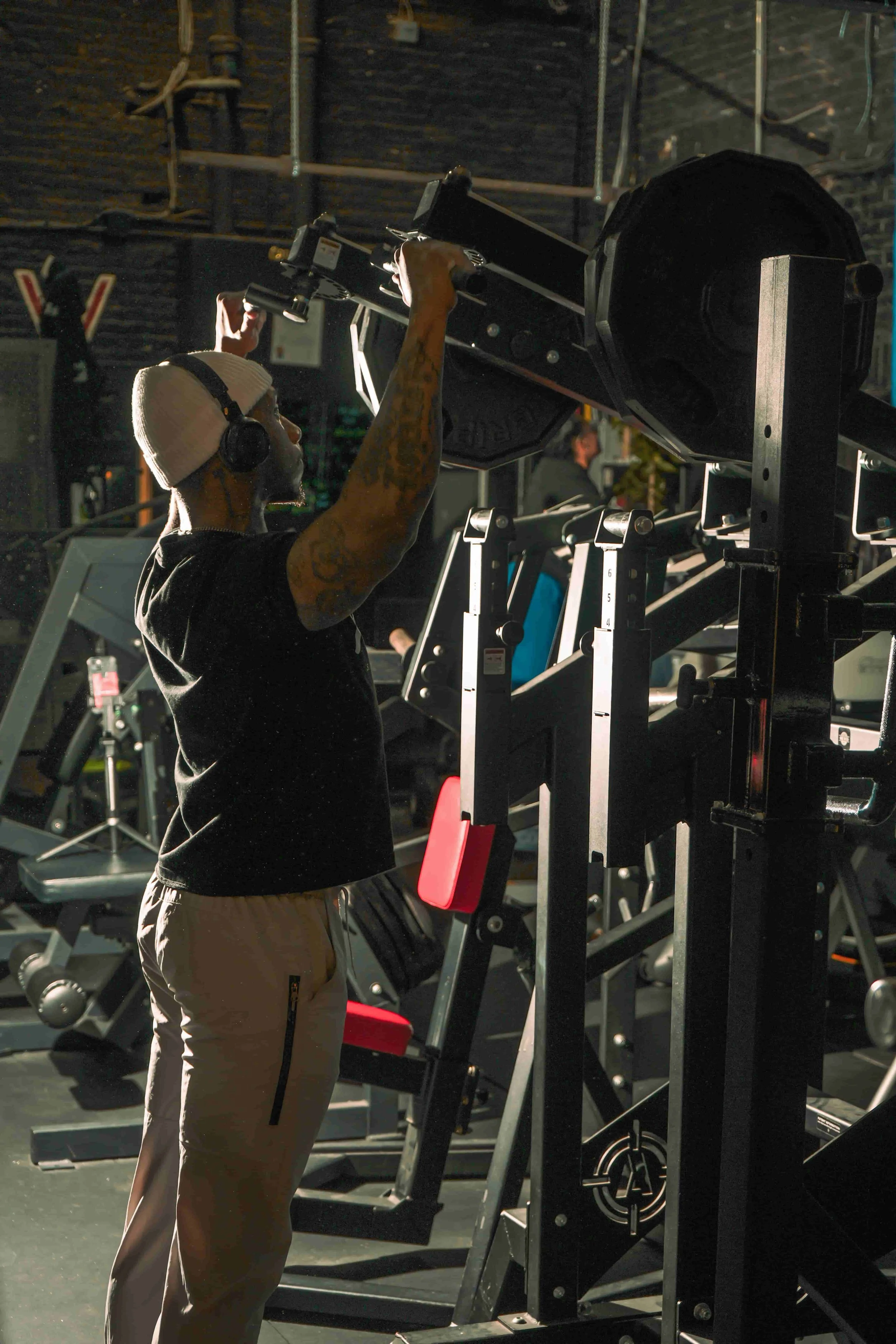 A man wearing a white beanie, black shirt, and beige pants is adjusting equipment at a gym. The gym has various exercise machines and gym equipment in the background.